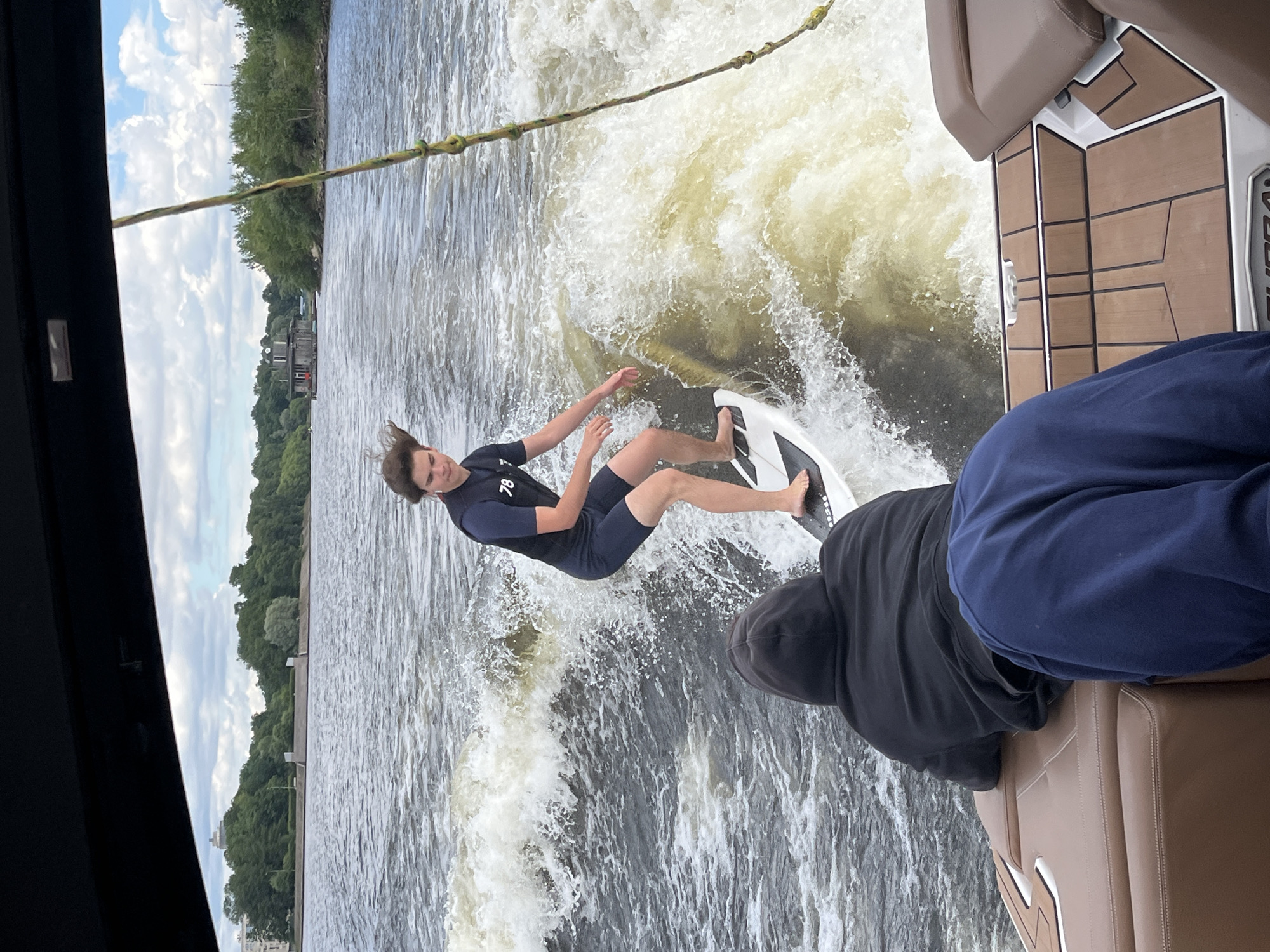 In this image, a young man is wakeboarding on a river. He is wearing a dark blue wetsuit with the number "78" visible on the back, and dark blue shorts. He is on a white wakeboard and appears to be in mid-air, performing a trick. The water is choppy and white with spray, indicating he is moving at speed behind a boat.
In the foreground, a person in a black hooded sweatshirt and navy blue pants is seated on a brown leather seat of a boat, partially obscuring the view of the wakeboarder. The boat has a wooden deck with textured brown panels.
The background shows a tree-lined shore with some buildings visible. The sky is partly cloudy. The image orientation is rotated, suggesting it was taken on a mobile device that was held vertically. The overall scene appears to be a recreational activity on a river, possibly during the daytime. The location is suggested to be Saint Petersburg, Russia.