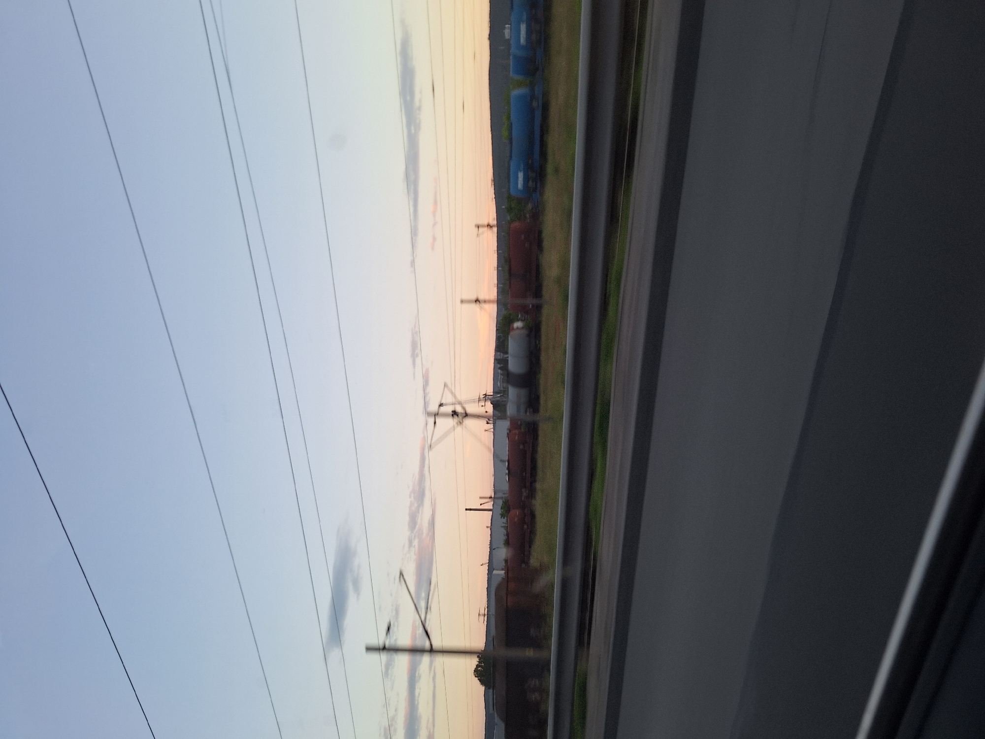 The image depicts an outdoor scene at twilight, captured from a moving vehicle in Varna, Bulgaria, overlooking a railway and industrial area.
In the immediate foreground, the bottom edge of the frame shows a dark, blurred section, likely part of a vehicle's interior (e.g., dashboard or window frame). Above this, a wide, dark grey band represents a road surface, exhibiting motion blur. A silver-grey metal guardrail runs horizontally across the lower mid-ground, separating the vantage point from the railway environment.
Beyond the guardrail, a strip of unkempt green and brown vegetation (grass, weeds) spans the scene. Parked on multiple parallel railway tracks within this area are various freight train cars. On the left, several dark reddish-brown boxcars or open wagons are visible. Further to the right, a series of cylindrical tank cars appear in different colors: reddish-brown, grey, and prominently, several bright blue tank cars with indistinct white markings or logos.
Interspersed throughout the railway area are several vertical utility poles, likely catenary poles supporting the overhead electrical lines that crisscross the sky. One prominent pole on the left shows clear structural details. In the middle distance, industrial structures are visible against the horizon, including at least one tall smokestack or chimney and a large gantry-style crane on the right.
The background consists of low, rolling hills covered in dark vegetation, establishing the horizon line. The sky above presents a gradient, transitioning from a pale, clear blue at the zenith to a warm, soft yellow and orange hue closer to the horizon, consistent with either sunrise or sunset. A few faint, wispy clouds are scattered in the lower sky. Numerous thin, dark overhead wires (presumably power lines or railway catenary) are strung across the entire upper portion of the sky.
No people are visible in the image. No distinct actions or interactions are occurring, aside from the implied motion of the photographer's vantage point. The scene captures an industrial-railway corridor on the periphery of Varna, Bulgaria, under soft twilight conditions.