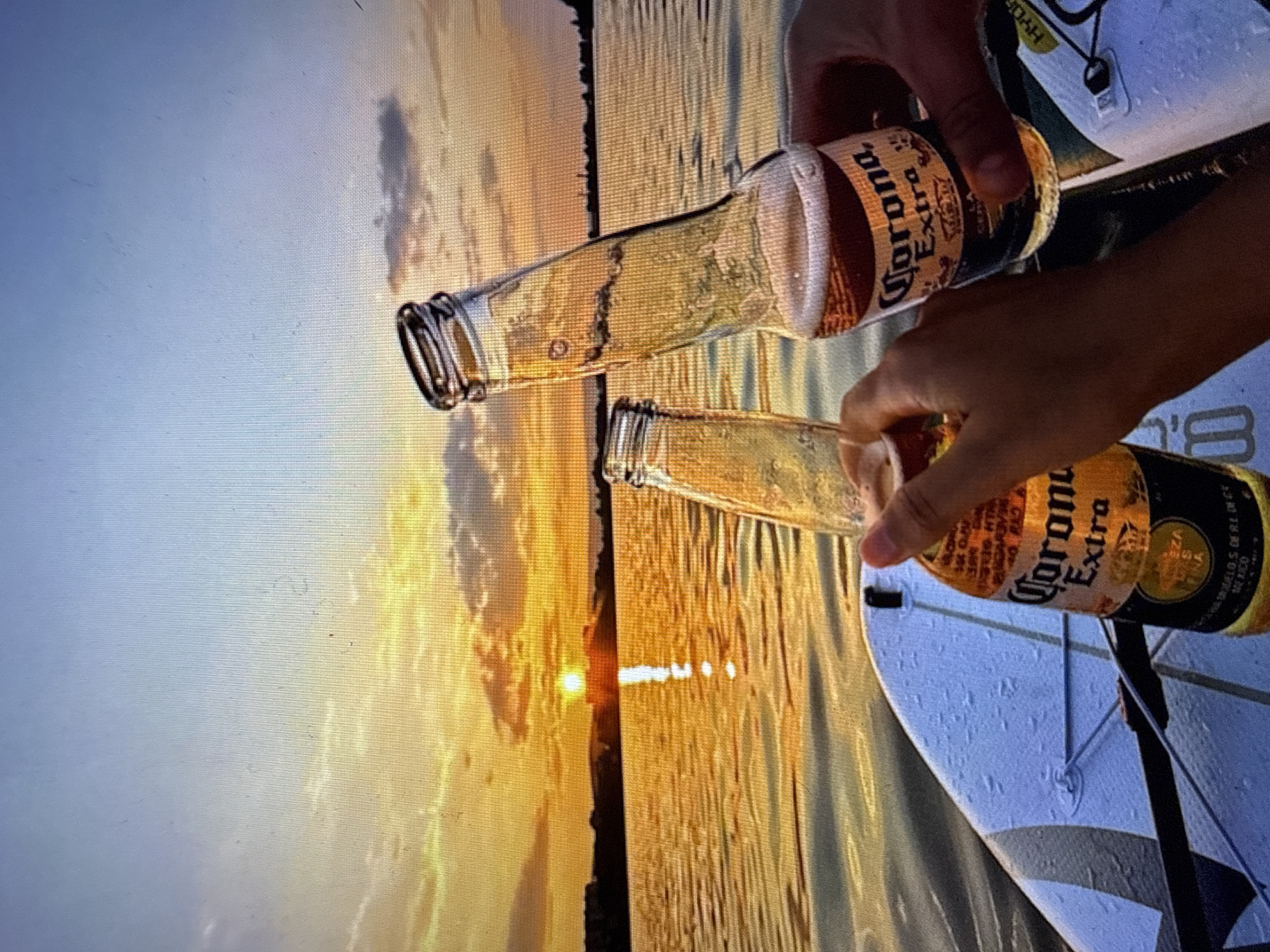 The image depicts a high-angle shot of two hands holding Corona Extra beer bottles over a paddleboard on a body of water at sunset, likely in the vicinity of Kardzhali, Bulgaria. The sky transitions from a light blue at the top to vibrant orange and yellow hues at the horizon, with scattered dark clouds visible, particularly in the lower sky and reflecting in the water. The sun, a bright orange orb, is visible directly above the distant shoreline, casting a prominent, shimmering reflection path across the calm water towards the viewer. The distant land appears as a dark, indistinct silhouette against the bright sunset, suggesting a tree line or small structures. In the foreground, the edge of a white and gray paddleboard is visible, textured and wet, with what appears to be part of a black paddle strap or bungee cord attached. Two hands, one on the left and one on the right, hold clear glass bottles of Corona Extra beer. The bottle on the left is mostly full, while the bottle on the right shows a partially consumed beverage with a frothy head, indicating recent drinking. The bottles are tilted slightly, seemingly being brought together for a toast or shared moment. The entire image exhibits a visible grid pattern, suggesting it is a photograph of a digital screen or a heavily processed digital image.