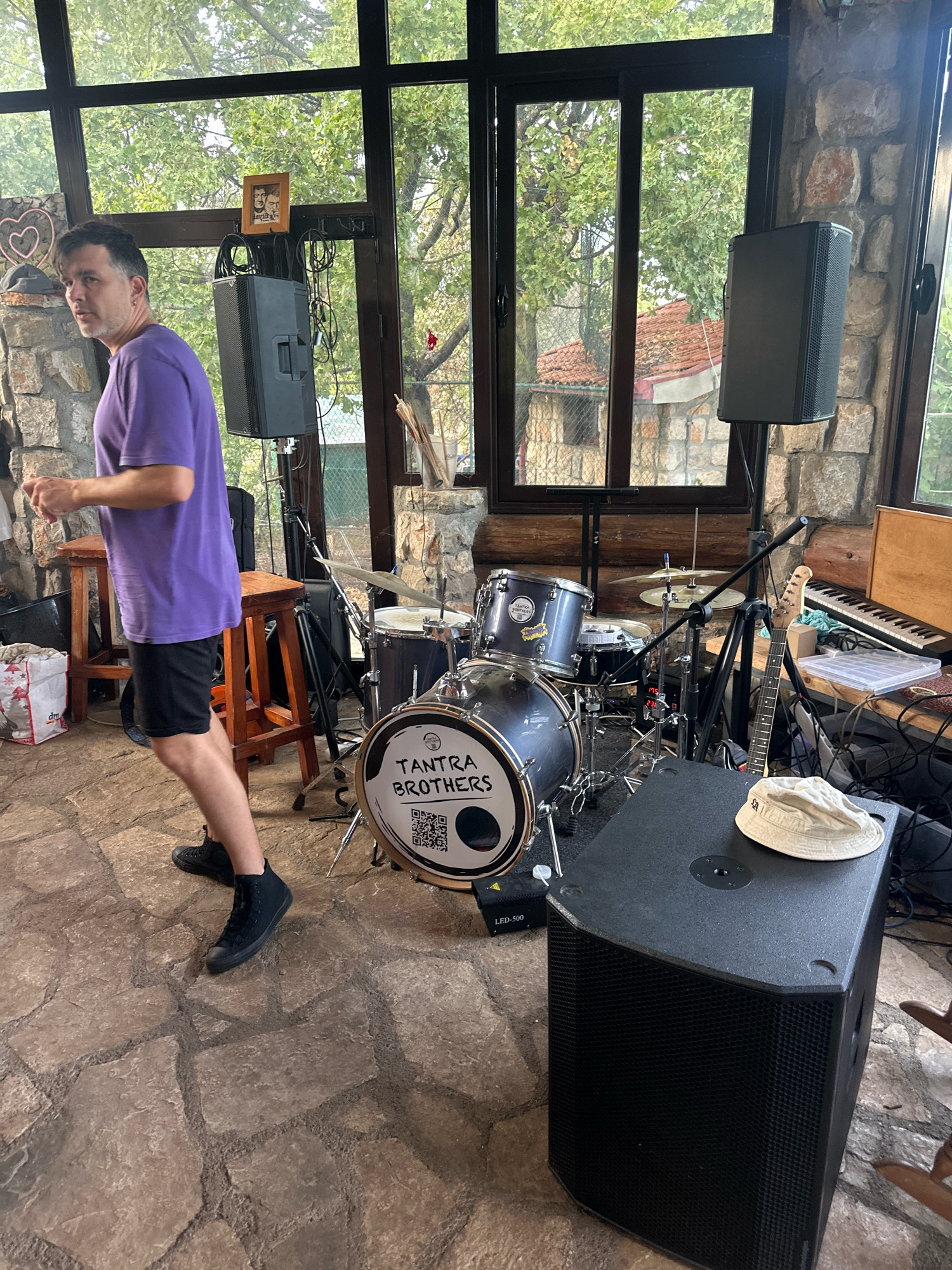 A man in a purple t-shirt and black shorts is standing in what appears to be a room with large windows overlooking trees and buildings. The room has a stone wall and a stone floor. In the background, there is a drum set with "TANTRA BROTHERS" written on the bass drum, a guitar, and a keyboard. There are also speakers and other audio equipment. The man is looking to his left and appears to be in motion, possibly dancing or preparing to perform. The overall impression is of a performance space, likely a venue in Cetinje, Montenegro, given the context.