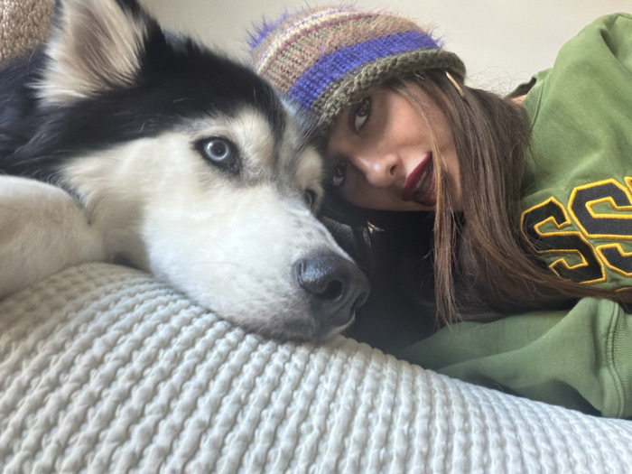 An indoor scene in Tel-Aviv, Israel, depicts a young female and a husky dog in close proximity. The dog, positioned on the left, is a black and white husky with prominent blue eyes, resting its head on a light-colored, ribbed knitted surface, possibly a pillow or blanket. The female, positioned on the right and slightly behind the dog, has long dark hair, wears dark red lipstick, and is dressed in a dark green sweatshirt featuring yellow/gold letters on the chest. She also wears a multi-colored knitted beanie with stripes of purple, brown, and green. Her head is tilted, and she gazes upwards and to the right. Both subjects appear relaxed, with their heads close together.