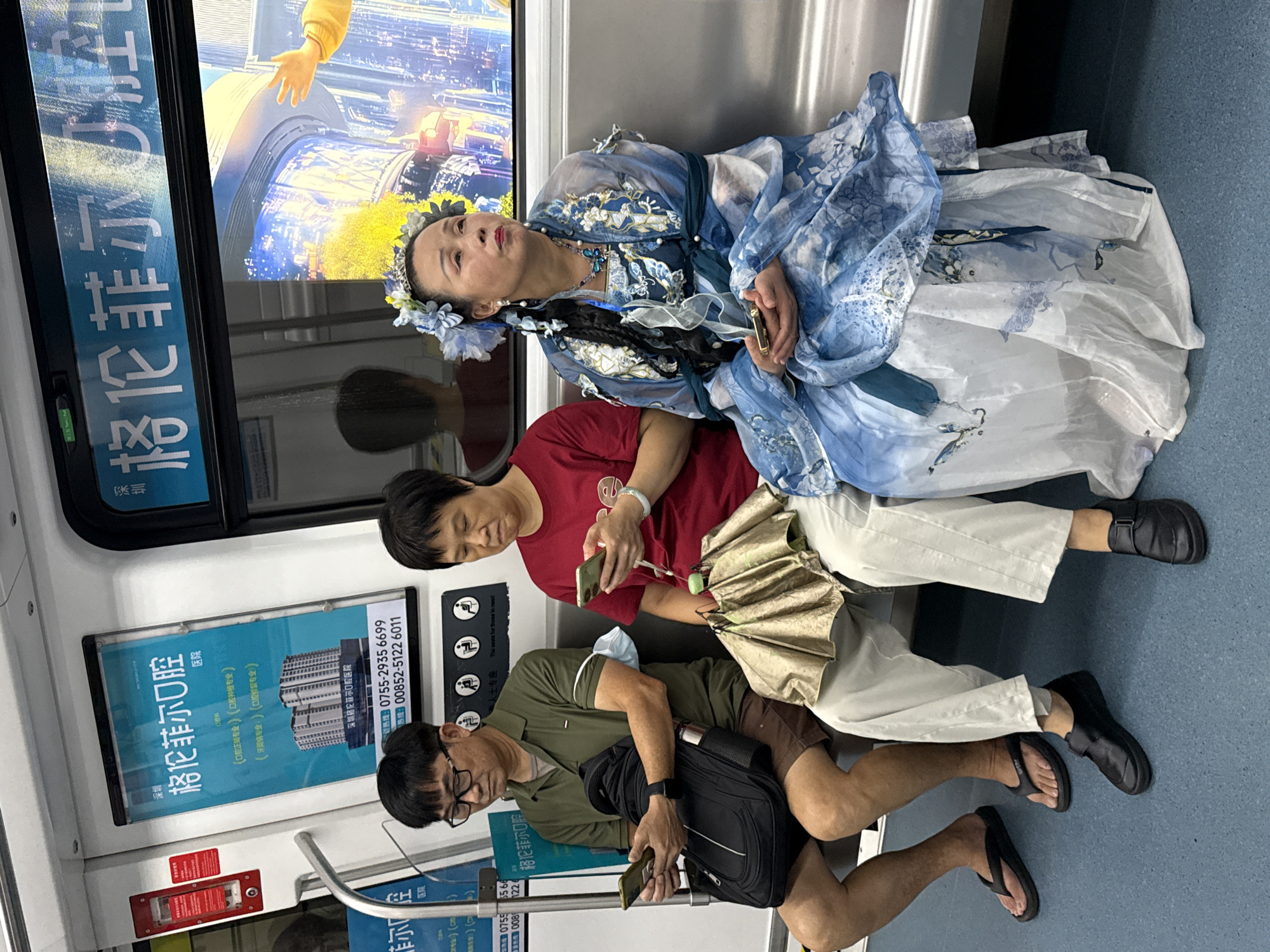 Three individuals are seated on a public transit vehicle, likely a subway train, with a backdrop of advertisements and cityscape imagery. The person on the left, a male with glasses, wears a green polo shirt and brown shorts, holding a smartphone in his left hand and a black backpack on his lap. To his right, a woman in a red t-shirt and white pants holds a smartphone, with a golden umbrella resting on her lap. The person on the far right, a woman adorned in a blue and white ornate traditional Chinese-style dress with floral hair accessories, gazes upward while holding a smartphone. The environment is a clean, modern train interior, with overhead advertisements and a digital display showing a vibrant urban scene, suggesting a location within Futian District, China.