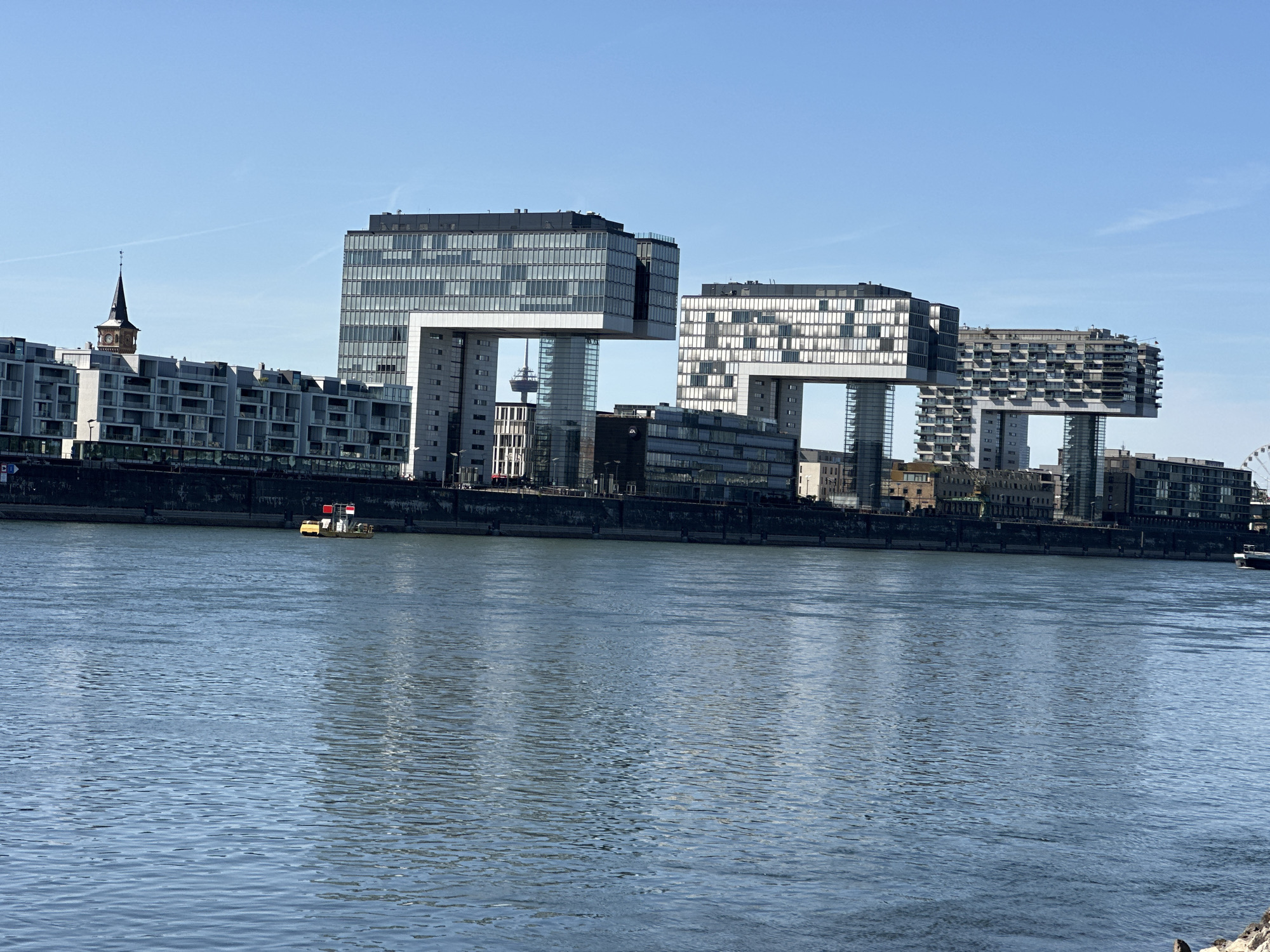The image shows the Kranhäuser, a distinctive architectural landmark in the city of Cologne, Germany, located on the Rhine river. The buildings are characterized by their C-shaped structure, with two buildings appearing to "lean" towards each other, creating a dramatic visual effect. In the foreground, the blue waters of the Rhine river are visible, with ripples on the surface. A small yellow boat with red flags is moving on the river near the embankment. Behind the river, the modern Kranhäuser buildings dominate the skyline, interspersed with other contemporary architectural structures. To the left, a traditional church spire with a clock can be seen, offering a contrast between old and new architecture. The sky is clear and blue, suggesting a sunny day. The scene captures a contemporary urban landscape with a prominent waterfront.