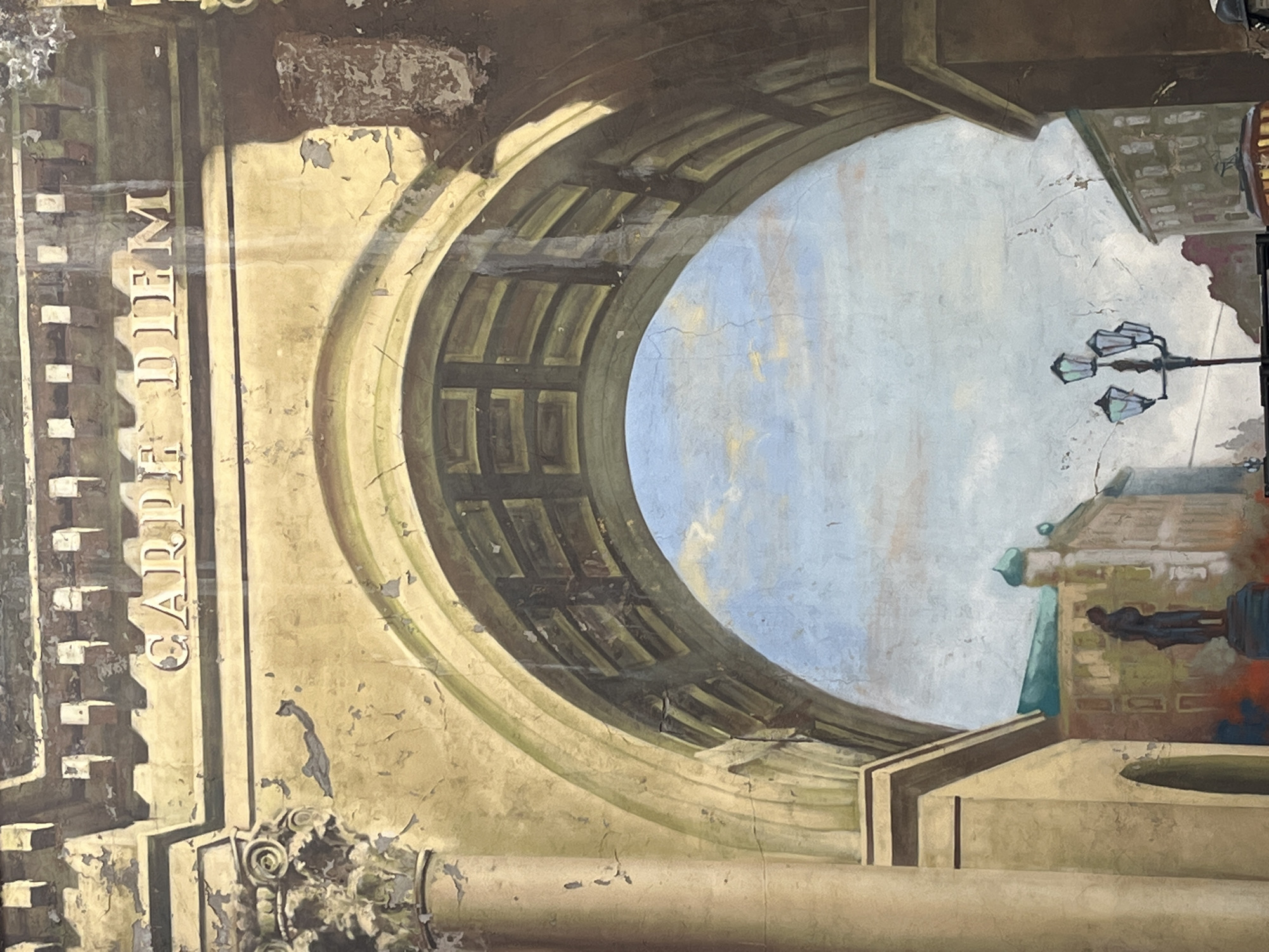 The image depicts a mural or painted scene, likely on a wall, showing an architectural structure with an arched opening. Through the arch, there's a view of a light blue sky with faint clouds, and a building exterior with street lamps and a statue. The word "CAPITOLIUM" is inscribed vertically on the left side of the structure. The image is likely depicting the Bolshoi Theatre in Moscow, Russia. The scene appears aged, with visible cracks and wear on the painted surface. The style is somewhat classical or neoclassical in its architectural details.
