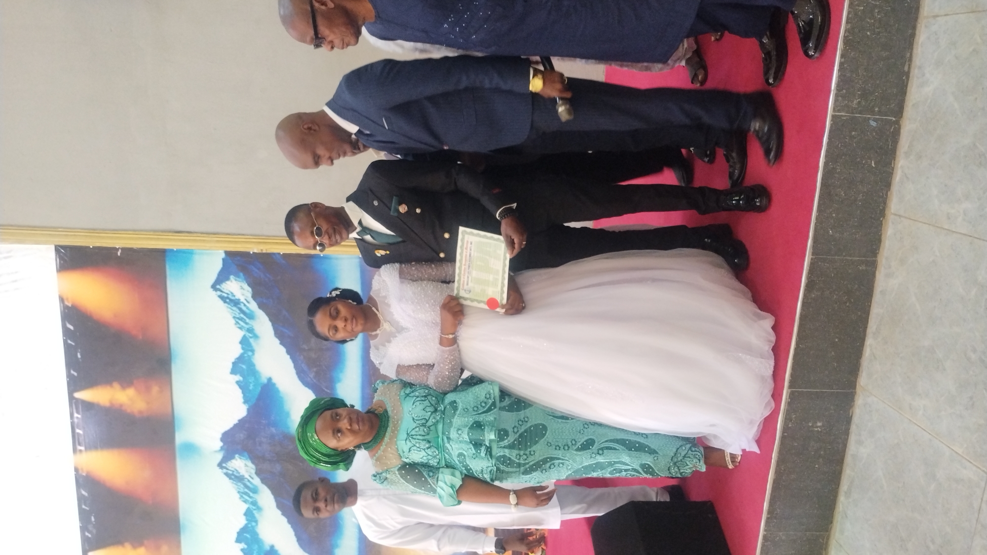 In the city of Kabong, Nigeria, on a raised platform covered with a red carpet, a wedding ceremony is taking place. A bride, wearing a white wedding gown, stands center, holding a certificate. To her left, a woman in a green patterned traditional outfit with a matching headwrap stands next to a man in a white garment. To the bride's right, a groom in a dark suit with a green tie and pocket square holds the certificate with her. Behind them, two men in suits observe the proceedings. The backdrop features a panoramic image of snow-capped mountains under a dramatic, fiery sky. The scene captures a moment of celebration and formal recognition of the union.