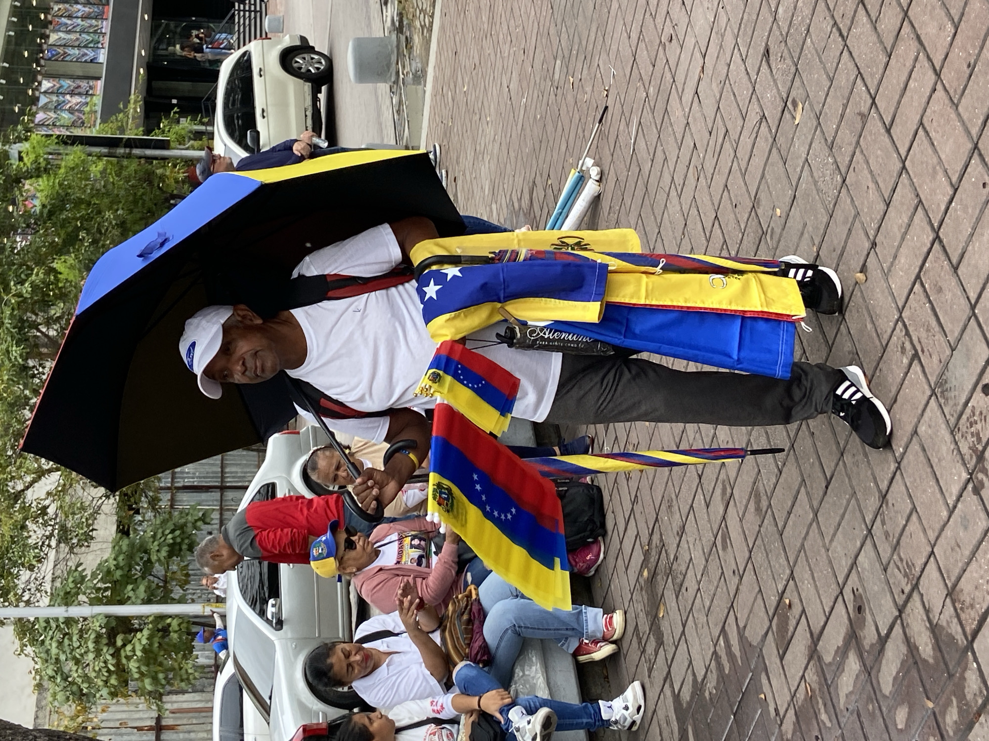 The image depicts a lively daytime street scene in Caracas, Venezuela, centered around a man who appears to be a vendor. He stands on a paved sidewalk, holding a large blue and black umbrella open above him. He is dressed in a white t-shirt, dark trousers, and black and white athletic shoes, topped with a white baseball cap featuring a white logo, possibly "FORD." He has a friendly expression, smiling towards the viewer.
The man is actively displaying and likely selling numerous Venezuelan flags, characterized by their prominent yellow, blue, and red stripes, eight white stars arranged in an arc, and the national coat of arms. Several flags are folded and draped over his arm, while others are unfurled, showcasing their vibrant colors. Alongside the flags, various colorful umbrellas, echoing the hues of the Venezuelan flag (yellow, blue, red), are either held or leaning against him. One rolled item near his leg features the visible text "Alenuiu" and "PARA GENTE COMO TU" (for people like you).
In the background, several other individuals are present; some are seated on a low wall or curb, while others are standing or walking. A white car is parked further back on the street, and commercial buildings line the background, indicating an urban setting. The sidewalk is paved with diamond-patterned blocks. The overall atmosphere suggests a public gathering or a busy commercial area.