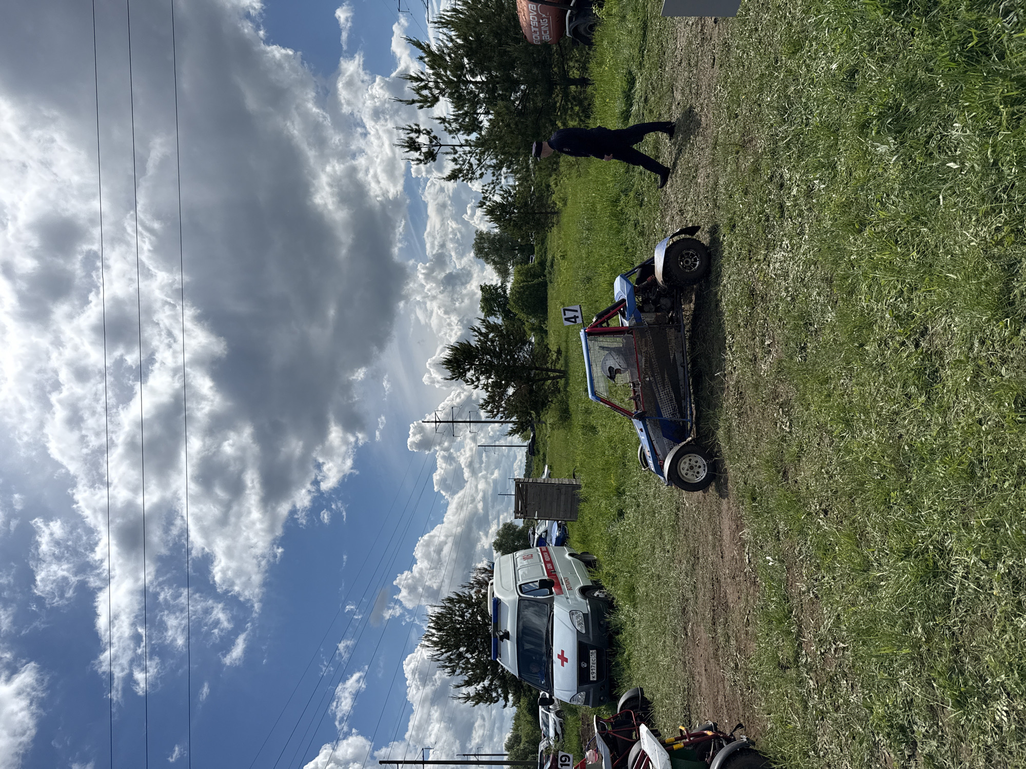 The image depicts an outdoor scene on a partly cloudy day, possibly in the city of Votkinsk, Russia, as there is an ambulance with a Russian license plate. The foreground is a grassy area with a dirt path. In the middle ground, there is a blue and red buggy with the number "47" on it. A man dressed in dark clothing is walking on the grass behind the buggy. To the left, there is an ambulance and other vehicles parked. Power lines are visible in the upper left corner, and trees and other foliage are scattered in the background.
