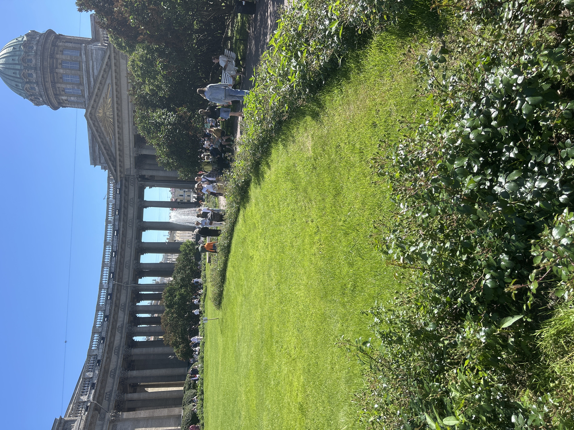 In an urban park setting, a curved colonnade with classical columns and an ornate balustrade forms the backdrop for a vibrant green lawn. In the distance, a large dome with a green patina rises above the structure, suggesting a significant architectural landmark. Several people are visible throughout the scene, with some seated on benches, others walking or standing in small groups on the grass. A fountain with a powerful spray of water is also present, adding to the lively atmosphere of the park. The foreground is dominated by lush green bushes, their leaves catching the bright sunlight. The sky above is a clear, bright blue.