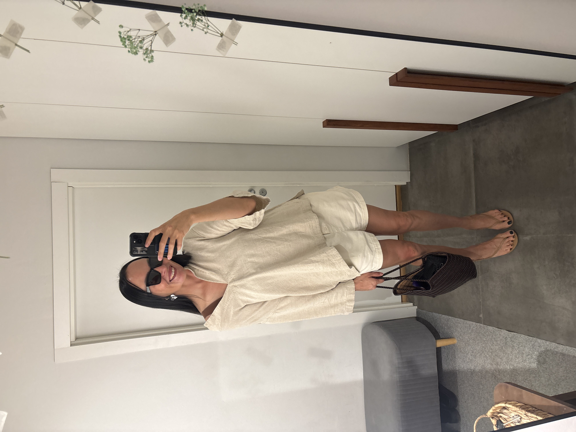 The image shows a woman taking a selfie in what appears to be a dressing room. She is wearing a light-colored linen shirt and shorts and sunglasses. She is holding a woven handbag. The dressing room has white walls and a gray floor. A gray padded bench is visible. The mirror has a black frame. The location is unknown, and therefore I cannot determine if it's in Chaikovsky, Russia.