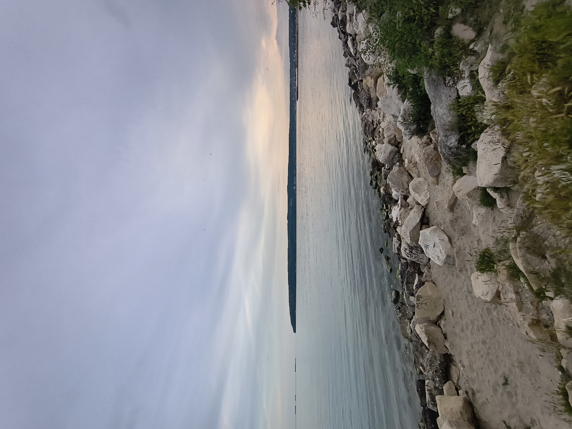 The image depicts a shoreline in Varna, Bulgaria. On the right, the shoreline consists of large rocks, sand, and vegetation. The water is on the right. On the left, the sky is predominantly cloudy with shades of gray and pale orange near the horizon. A thin strip of land is visible along the horizon. The scene is set at dusk or dawn.