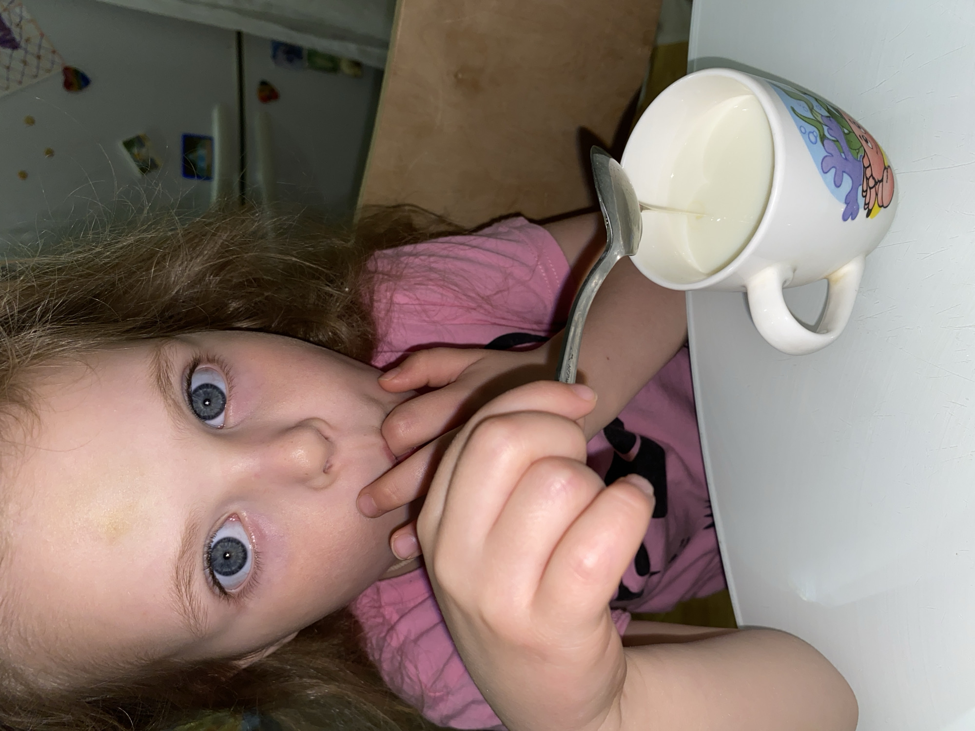 A close-up, portrait-oriented image shows a young child, likely a girl, with light brown or blond hair and striking, wide blue eyes, looking directly at the camera. Her face dominates the left side of the frame, and one of her fingers is held near her mouth. She is wearing a pink t-shirt with a dark design partially visible on the fabric.
To the child's right, a white ceramic mug, decorated with colorful cartoon-like sea creatures (a pink crab and blue/green coral/seaweed), sits on a clean white table surface. The mug contains a white liquid, presumably milk, and a silver spoon is partially immersed in it. The scene suggests the child is either about to drink, has just finished drinking, or is otherwise engaged with the mug of milk.
The background consists of a light brown wooden surface on the right, and a white appliance, possibly a refrigerator, on the left, adorned with several small, colorful magnets or drawings. The lighting is artificial and appears somewhat dim, indicating an indoor setting, likely a home kitchen or dining area. The child's expression is one of curiosity or mild surprise. There is no visible text in the image.