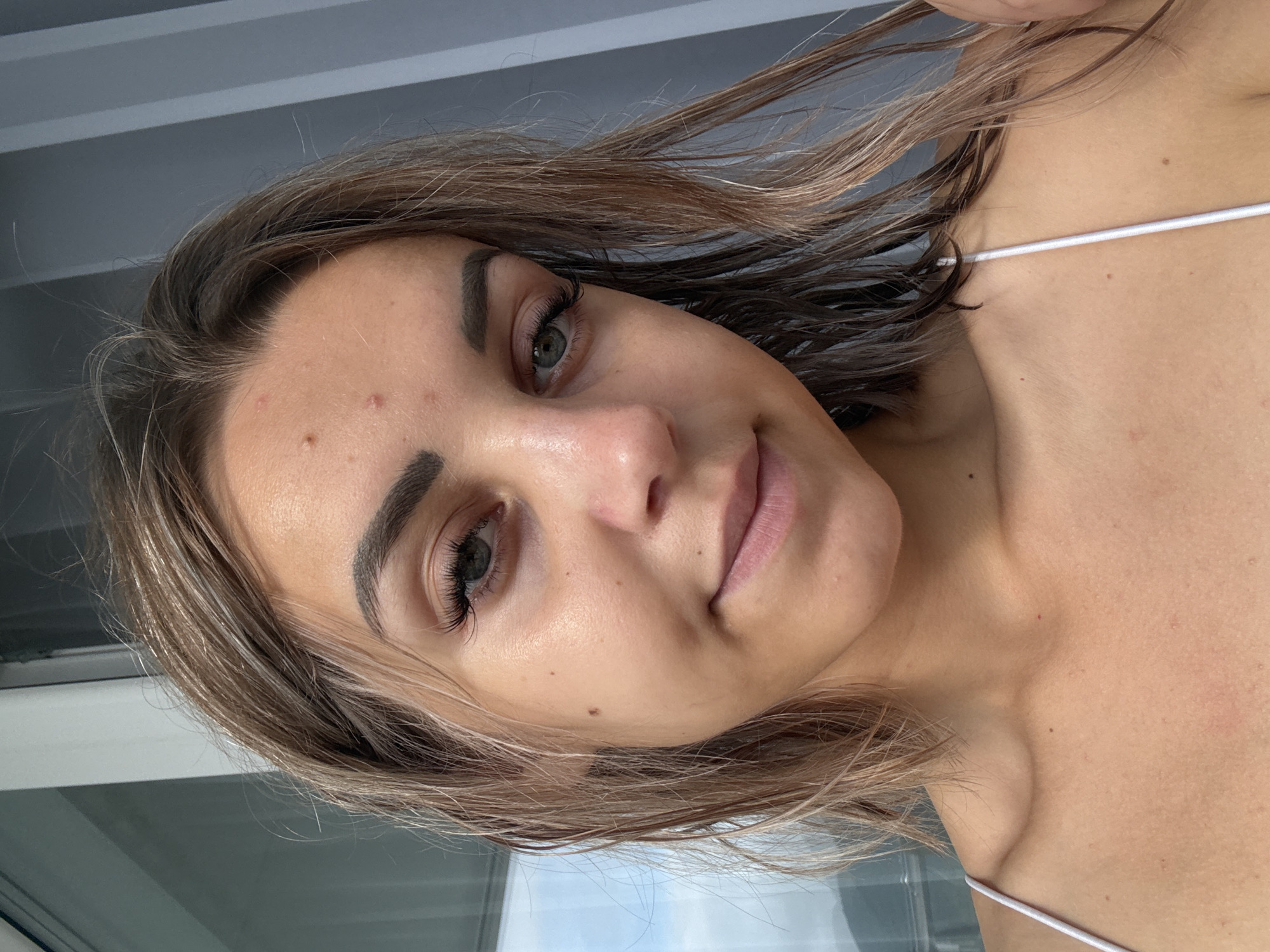 A close-up, front-facing, head-and-shoulders shot of a young woman with light brown, shoulder-length hair. She has fair skin with some blemishes on her forehead and cheek. Her eyes are a green-blue hue, and she is wearing false eyelashes. Her lips are slightly parted, and she has a subtle smile. She is wearing a white spaghetti strap top. The background appears to be an indoor setting with vertical blinds and a window showing a glimpse of the sky. The image does not contain any information that would allow for a precise location within Kholmsk, Russia.