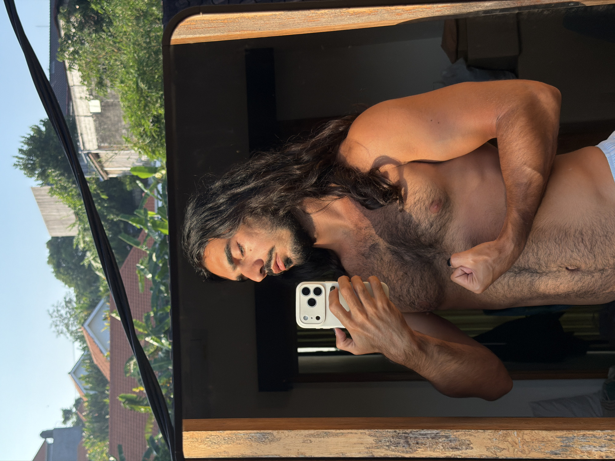 A man with long dark hair and a beard is reflected in a mirror. He is shirtless and wearing light blue underwear. He is holding a white smartphone in his right hand and flexing his left bicep. The background of the reflection shows houses with red-tiled roofs and lush green trees, indicating a tropical location. The overall lighting suggests it is late afternoon or early morning, with warm sunlight casting shadows. The man appears to be in a residential area in North Kuta, Indonesia, possibly on a balcony or porch.