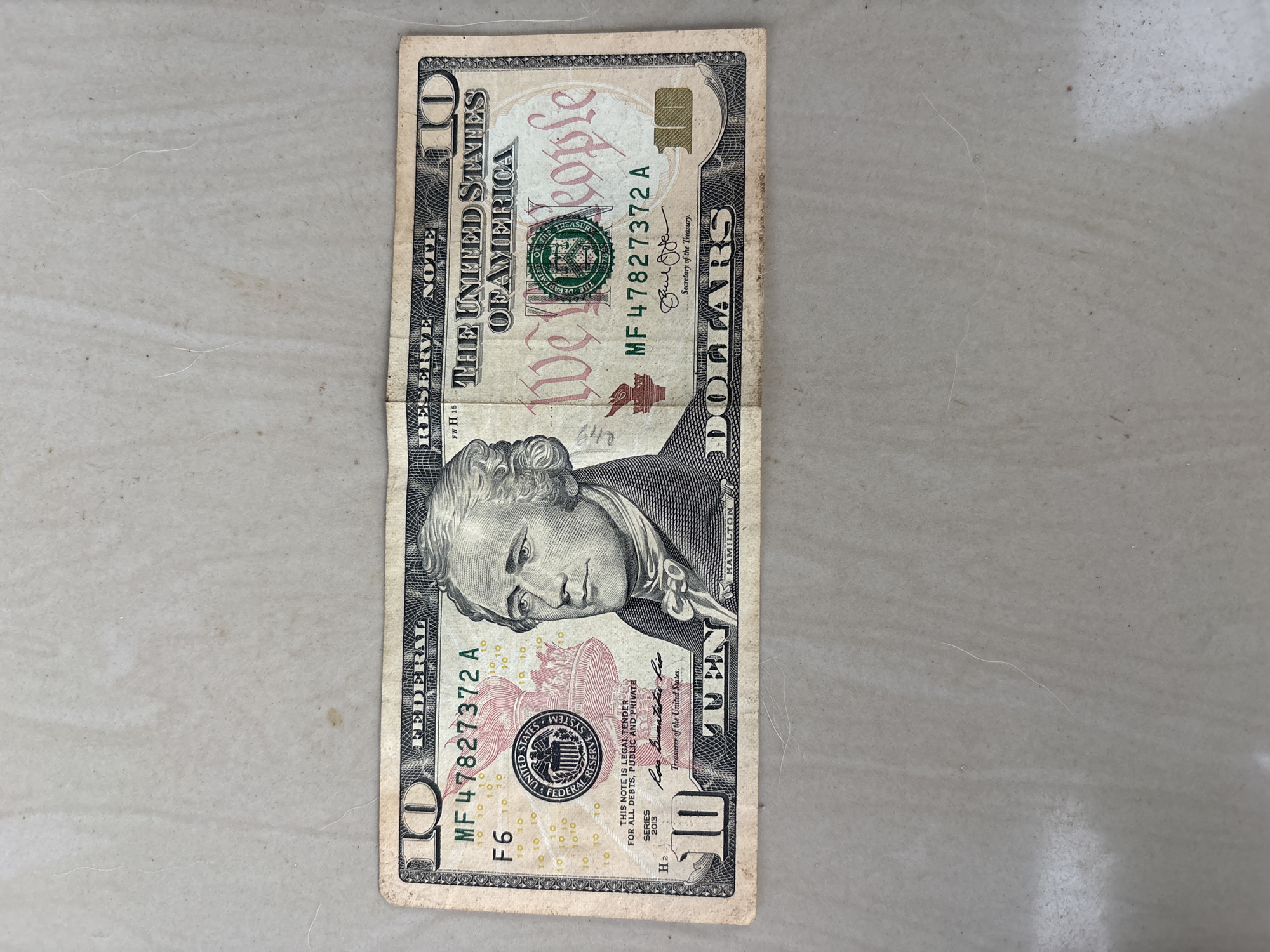 The image shows a slightly worn United States ten-dollar bill lying on a neutral-colored surface, likely a countertop or tile floor. The bill features the portrait of Alexander Hamilton and the text "THE UNITED STATES OF AMERICA." The serial number is MF 47827372 A. The bill appears to be an older version with some creasing. There is no information to suggest the image was taken in Ikot Ekpene, Nigeria.