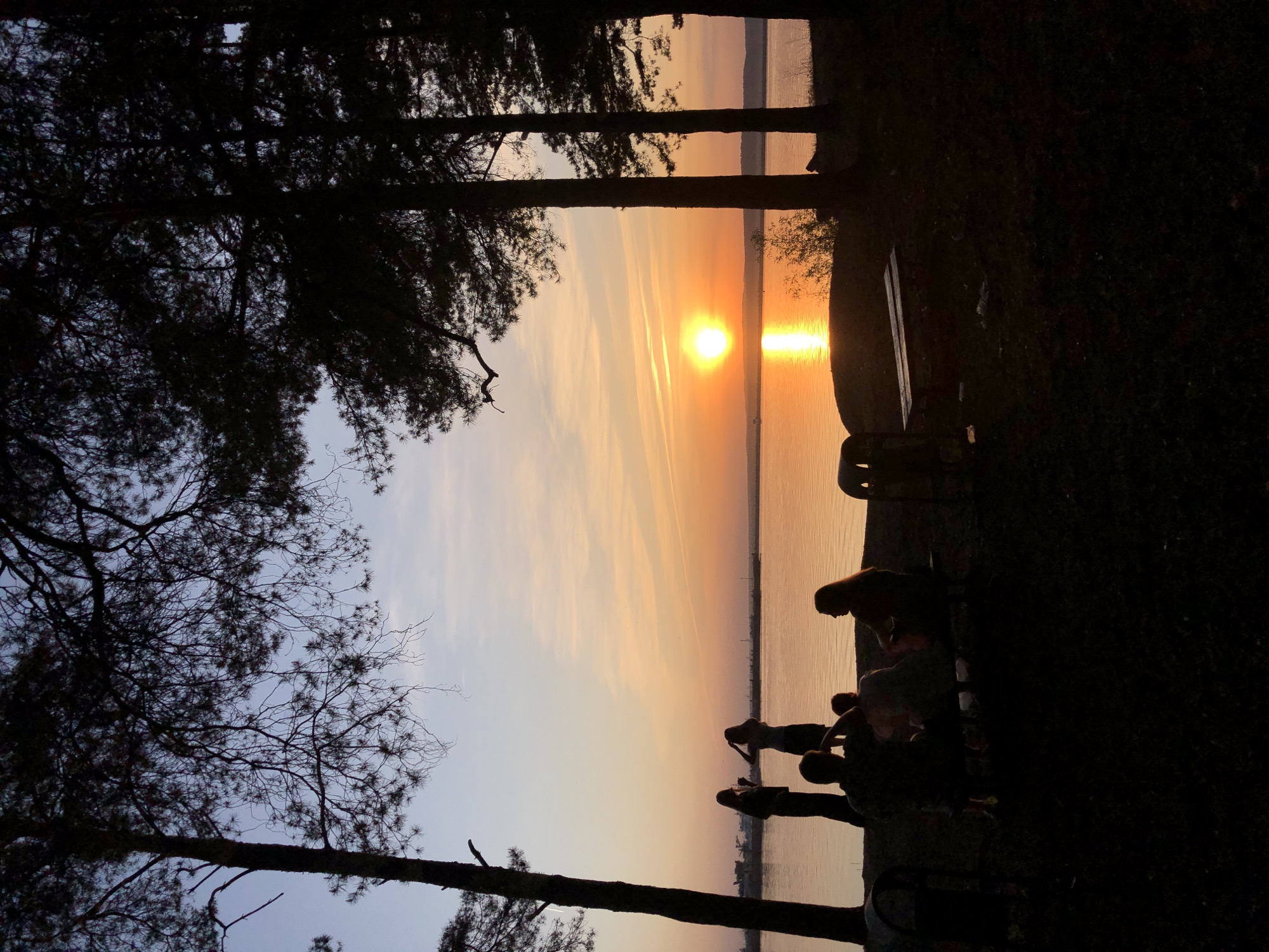 The image captures a sunset scene over a body of water, likely a lake or river, viewed from a location with trees. Silhouettes of people are visible on the right, some of whom appear to be standing or sitting. A bench is present near the people. The sun is setting directly over the water, creating a bright reflection. Trees frame the view on the left and top of the image. The image appears to be taken in Chaikovsky, Russia.
