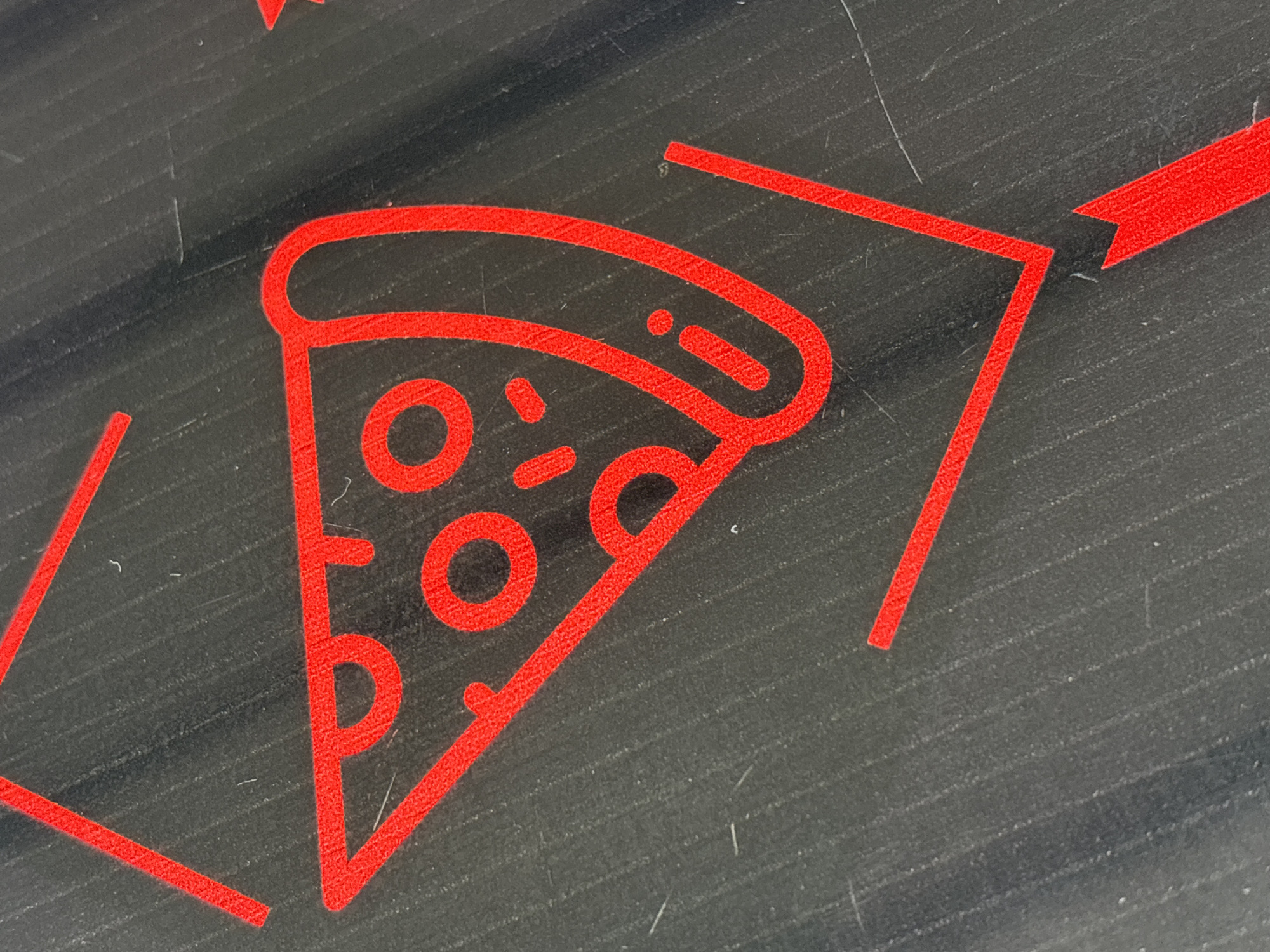 The image displays a close-up view of a red outline graphic depicting a slice of pizza, set against a dark, textured background. The pizza slice is minimalist in design, showing its crust, a curved top edge, and several circular and oval shapes representing toppings like pepperoni or olives. Surrounding the pizza icon are other red outline graphics, including straight lines and a right-pointing arrow, suggesting a directional or decorative element.
The background surface is predominantly dark gray or black, featuring subtle horizontal striations and numerous fine scratches, scuffs, and small specks of dust, indicating a worn or used material. The red color of the graphics stands out vibrantly against this muted background. No people or specific activity are visible. The image captures a static scene, likely a floor decal, a table design, or a sign. While the location context is Plovdiv, Bulgaria, the image itself does not provide explicit visual cues to this specific city. There is no visible text in the image.