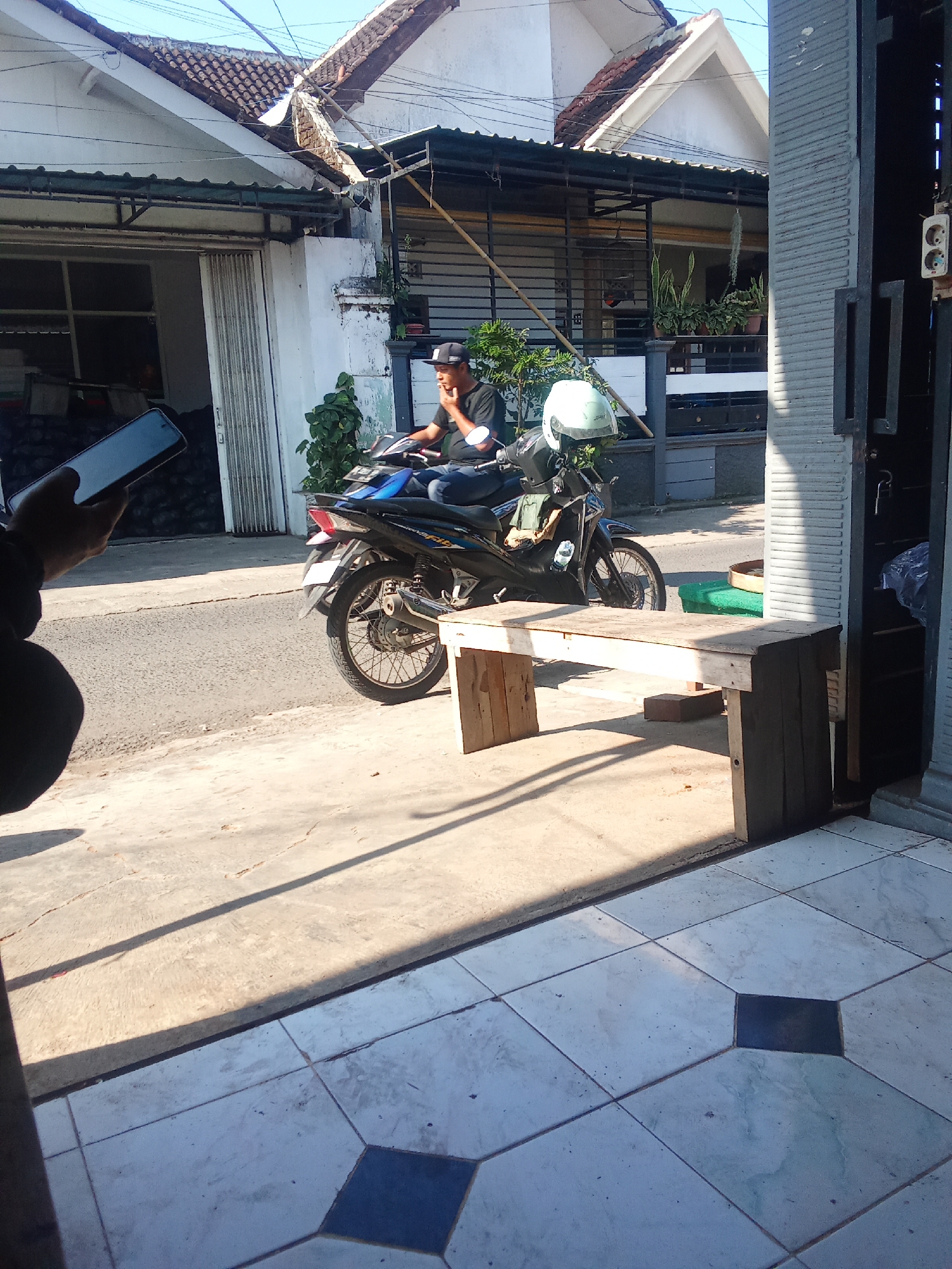 In Gentengwetan, Indonesia, a man is seated on a motorbike parked on a paved street in front of a building. The man wears a black t-shirt and a black baseball cap. He is looking away from the camera and appears to be holding a cigarette. The motorbike is blue and black, with a white helmet resting on the handlebars. In the foreground, a simple wooden bench is placed on the paved ground. The background shows a white building with a brown tiled roof. Sunlight casts long shadows across the scene. To the left, a person's hand is visible holding a smartphone.
