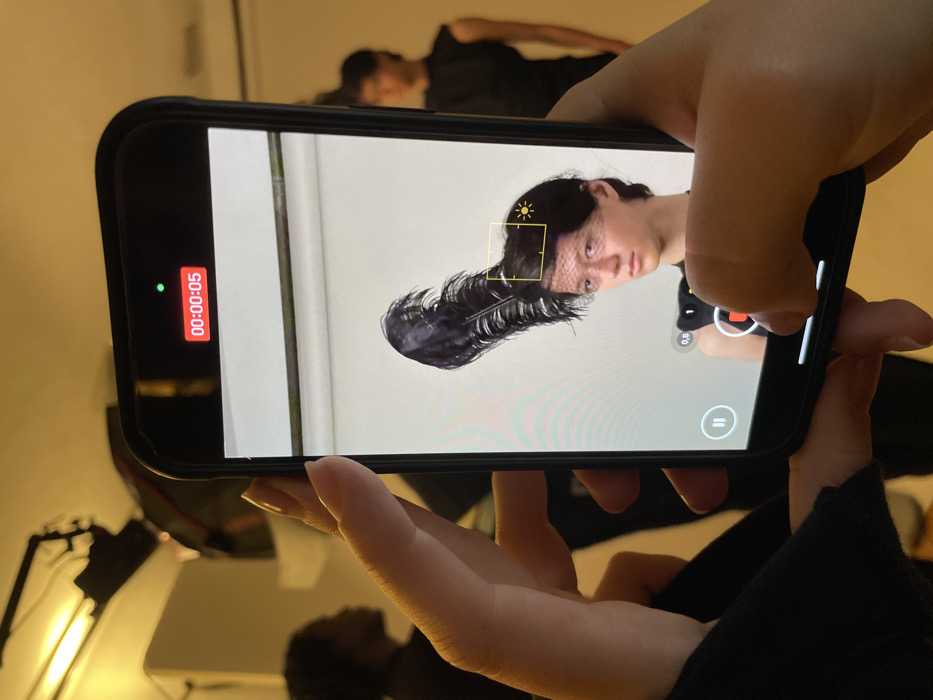 A person films a portrait of someone with a black feathered hairpiece on their phone. The subject on the phone's screen has fair skin. The phone is held horizontally in a person's hands. The phone's recording feature is active, as indicated by the green dot and red timestamp "00:00:05". In the background, another person is partially visible lying on the floor, and a softbox lamp stands nearby. The image captures the act of documenting or creating content.