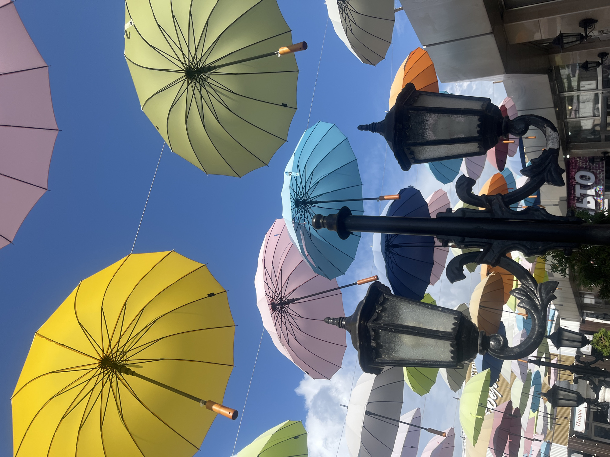 An outdoor scene features a vibrant canopy of colorful umbrellas suspended against a bright blue sky with scattered clouds. The umbrellas are in various shades of yellow, pink, light green, blue, and orange. Several black, ornate lampposts with glass panes are positioned in the foreground, some attached to buildings and others free-standing. In the background, the facade of a building with a striped pattern is visible, along with signs indicating businesses, including one with Cyrillic text that reads "ПИЦЦА" (PIZZA). The overall impression is a decorative street installation, likely in a pedestrian area or public space within the city of Sochi, Russia, designed to enhance the visual appeal of the urban environment. There are no people clearly visible in the image.