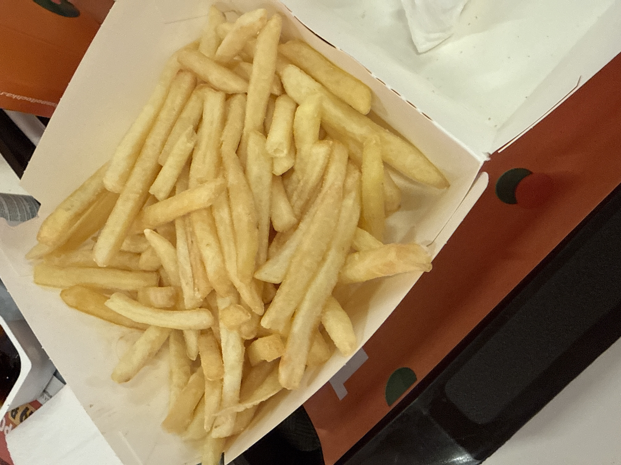 A cardboard container, full of french fries, sits on a table. Adjacent to the fries is another cardboard box. The setting appears to be indoors. The location is not discernible from the image.