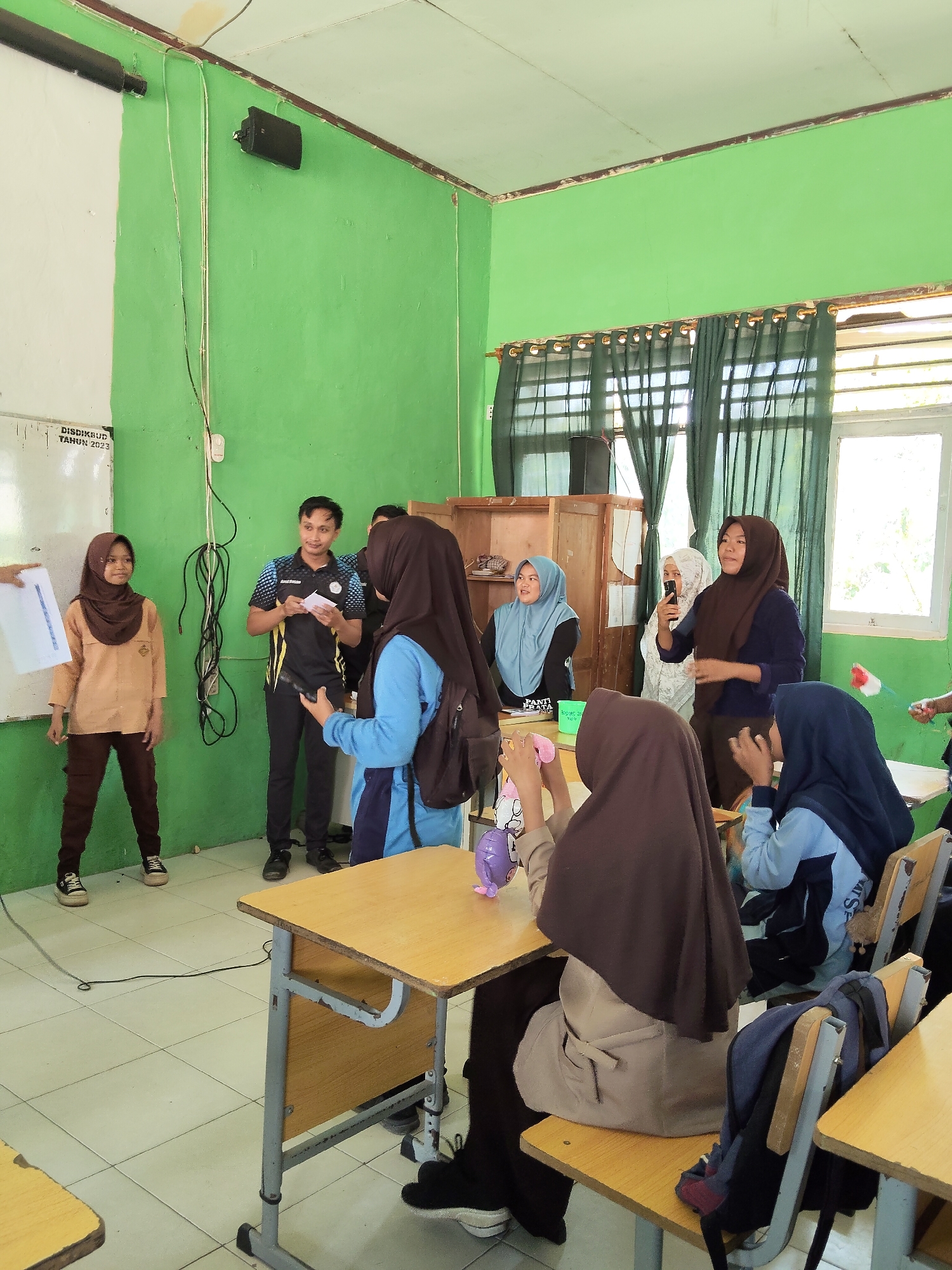 A group of students and an instructor are gathered in a classroom with bright green walls. A male instructor, wearing a blue and black polo shirt and black pants, stands in the center of the room holding a piece of paper. He appears to be addressing a group of students, some of whom are wearing hijabs. To his left, a female student in a tan uniform and brown hijab stands holding a piece of paper. To his right, another female student in a blue jacket and black hijab is facing him, and a few other students are seated at desks. The room contains typical classroom furniture, including desks, chairs, a whiteboard, and a projector screen. The overall impression is a school setting in Indonesia, likely a classroom for young students.