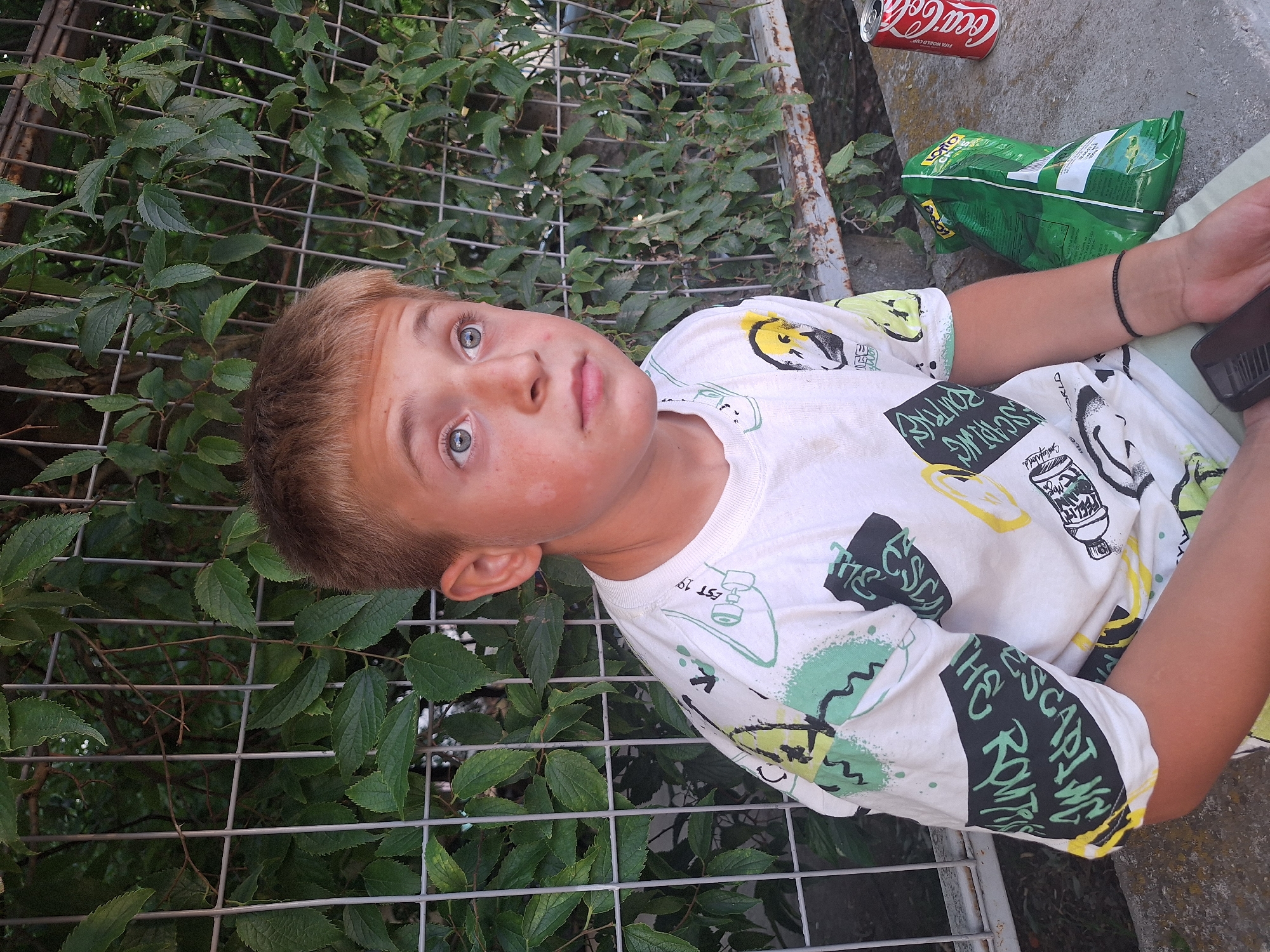 A young boy with blond hair and blue eyes, wearing a white t-shirt with a colorful graphic print, sits outdoors. He is looking off to the right with a neutral expression. Behind him is a metal fence covered in green foliage. To his right, on the ground, are a bag of chips and a can of Coca-Cola. The scene appears to be in Yambol, Bulgaria.