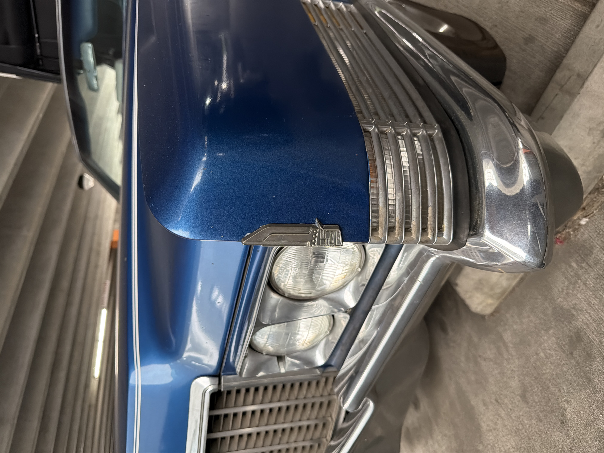 This is a close-up, eye-level, vertical, outdoor, daytime photograph of the front fender and grille of a vintage blue Cadillac. The car is parked in a concrete parking garage. The car has a shiny blue paint job with thin white pinstripes along the top of the fender. The front of the car features a chrome grille and two large, round headlights. A chrome Cadillac emblem is visible on the fender. The background shows the ribbed concrete ceiling of the parking garage. The overall condition of the car appears to be good, though there are some minor scratches and dust visible on the paint.