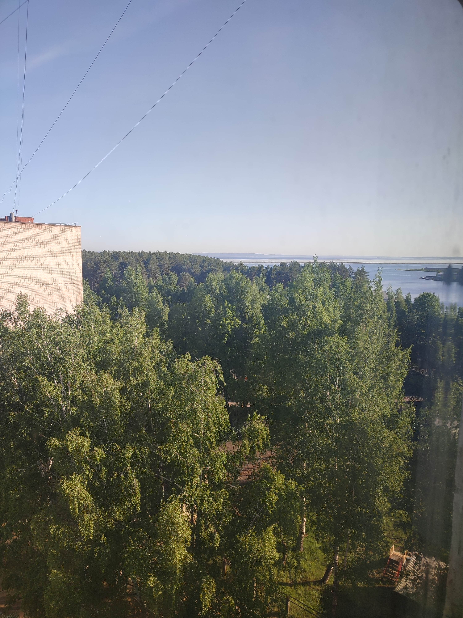 A slightly high-angle view of a landscape in the city of Chaikovsky, Russia. The sky is bright blue, fading to white near the horizon. A building with beige brick facade is on the left, partly obscured by lush green trees. A dense forest covers the middle ground, transitioning to a body of water in the background. Power lines stretch across the upper portion of the frame. Below the trees, a small grassy area with a wooden fence can be seen, along with a possible structure made of red and silver materials. The image appears to be taken from indoors, possibly through a window, given the faint reflection in the lower right corner.