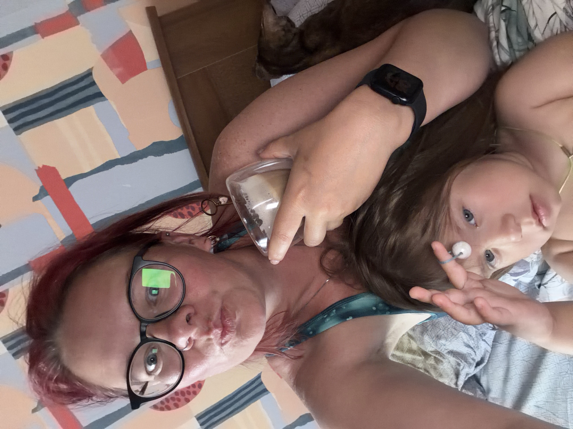 A selfie shows a woman and a young child lying down. The woman has red hair, glasses, and a nose ring. She is holding a glass containing a white substance. The child is beside her. Both are on a bed with patterned linens. A cat is visible in the background. The location is Unknown, Unknown.