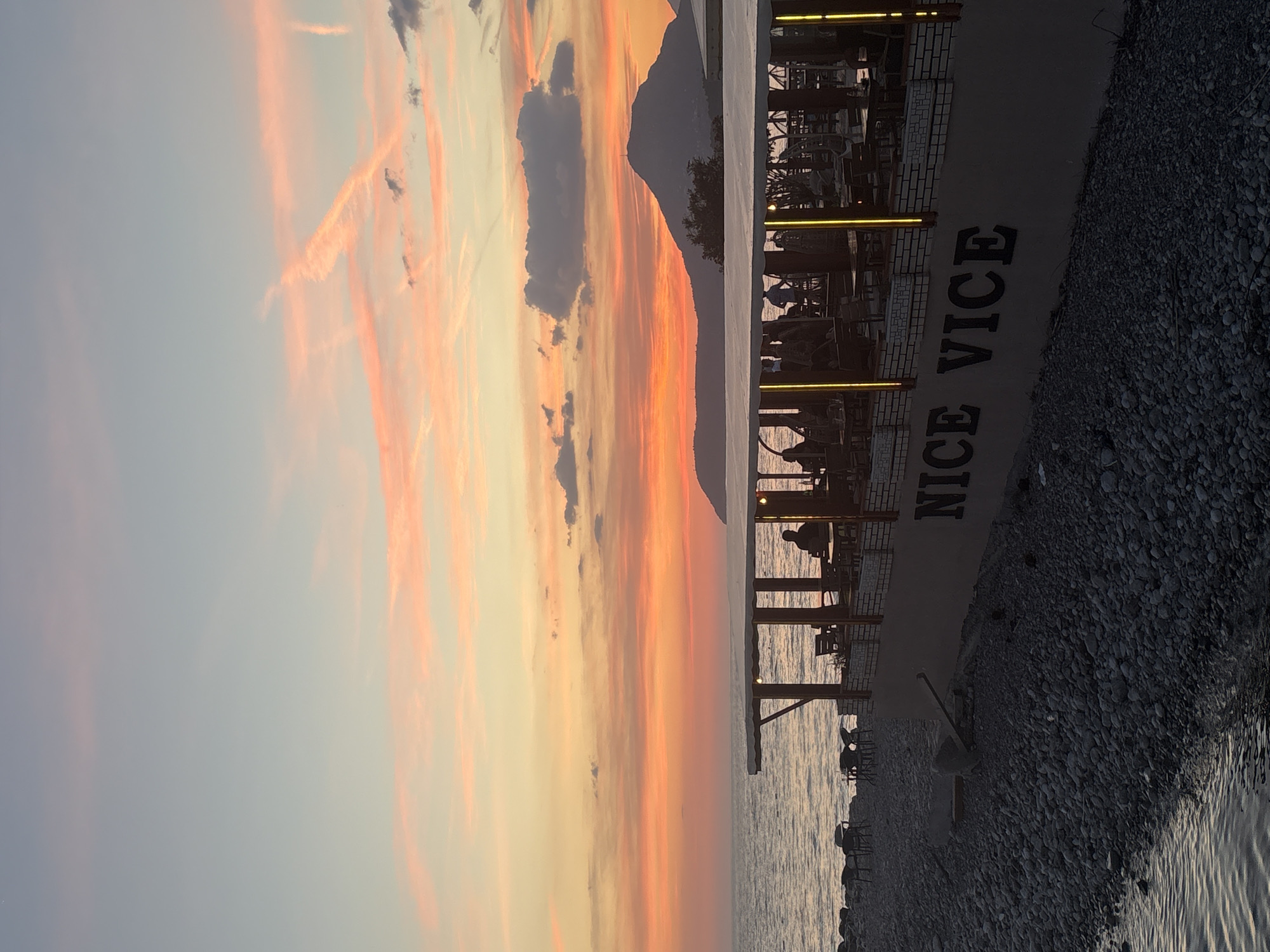 The image depicts a sunset scene in Bar, Montenegro. The sky displays a gradient of colors, transitioning from light blue to orange and pink, with streaks of clouds. The foreground features a pier or boardwalk with outdoor seating, possibly a restaurant or cafe named "NICE VICE," with the name vertically written on the side of the structure. People are seated at some of the tables, visible in silhouette. The pier extends over the water, which reflects the colors of the sky. The edge of the beach is visible, covered with pebbles or small rocks. A mountain silhouette is present in the background.