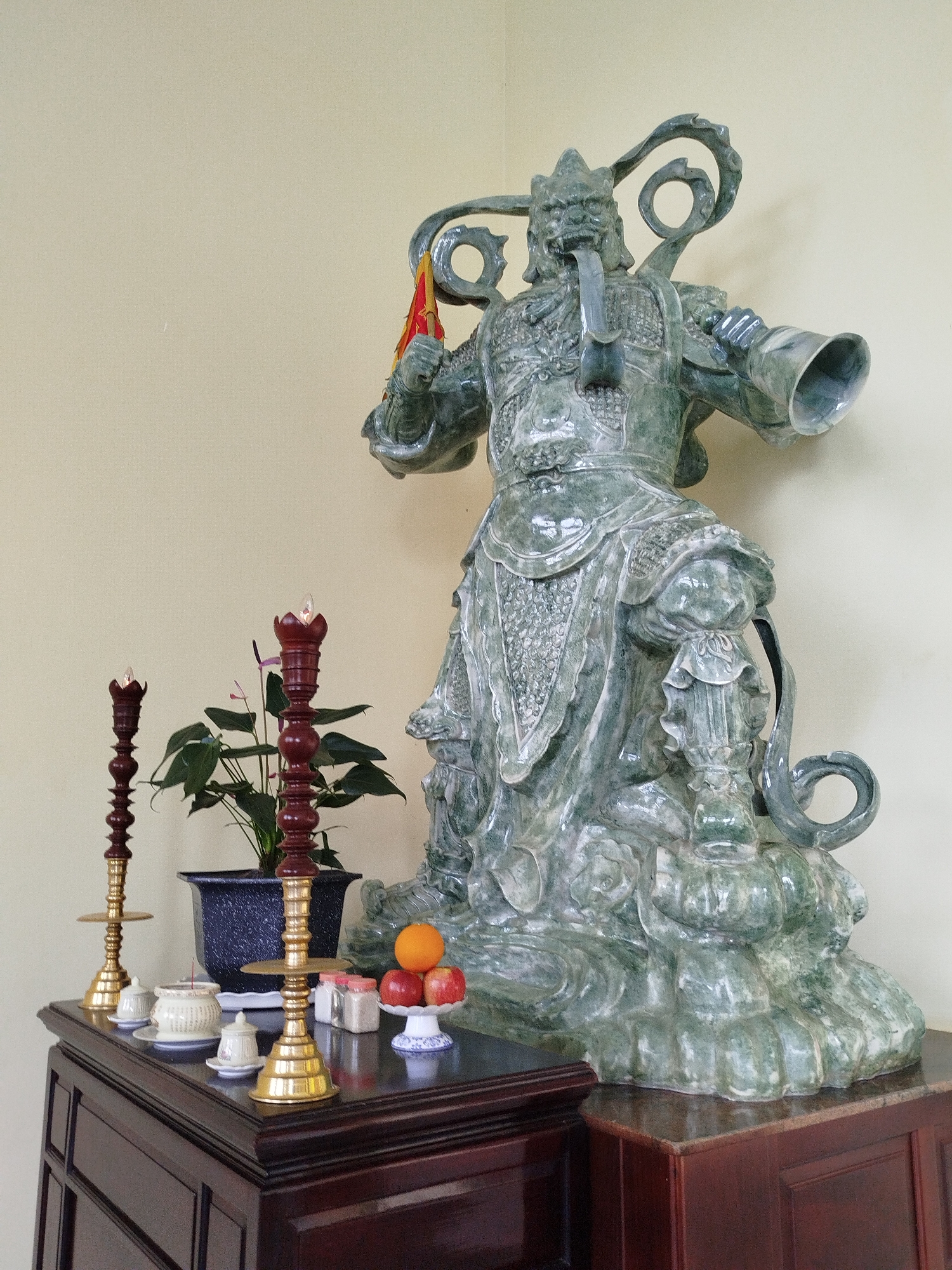 The image depicts a shrine in Lang Biang - Da Lat, Vietnam. On a dark wooden table is a large green statue of a deity, positioned to the right. It has ornate headgear and holds a small red and yellow flag. To the left of the statue is a potted plant with green leaves and red flowers. In front of the plant are two tall, ornate candleholders with unlit electric candles at the top. Between the candleholders and the statue is a small assortment of offerings including small white containers, a dish with incense, and a dish holding an orange and three red apples. The background is a plain, light yellow wall. The furniture is a rich, polished brown wood. The statue is sitting on top of another piece of furniture, also made of wood.