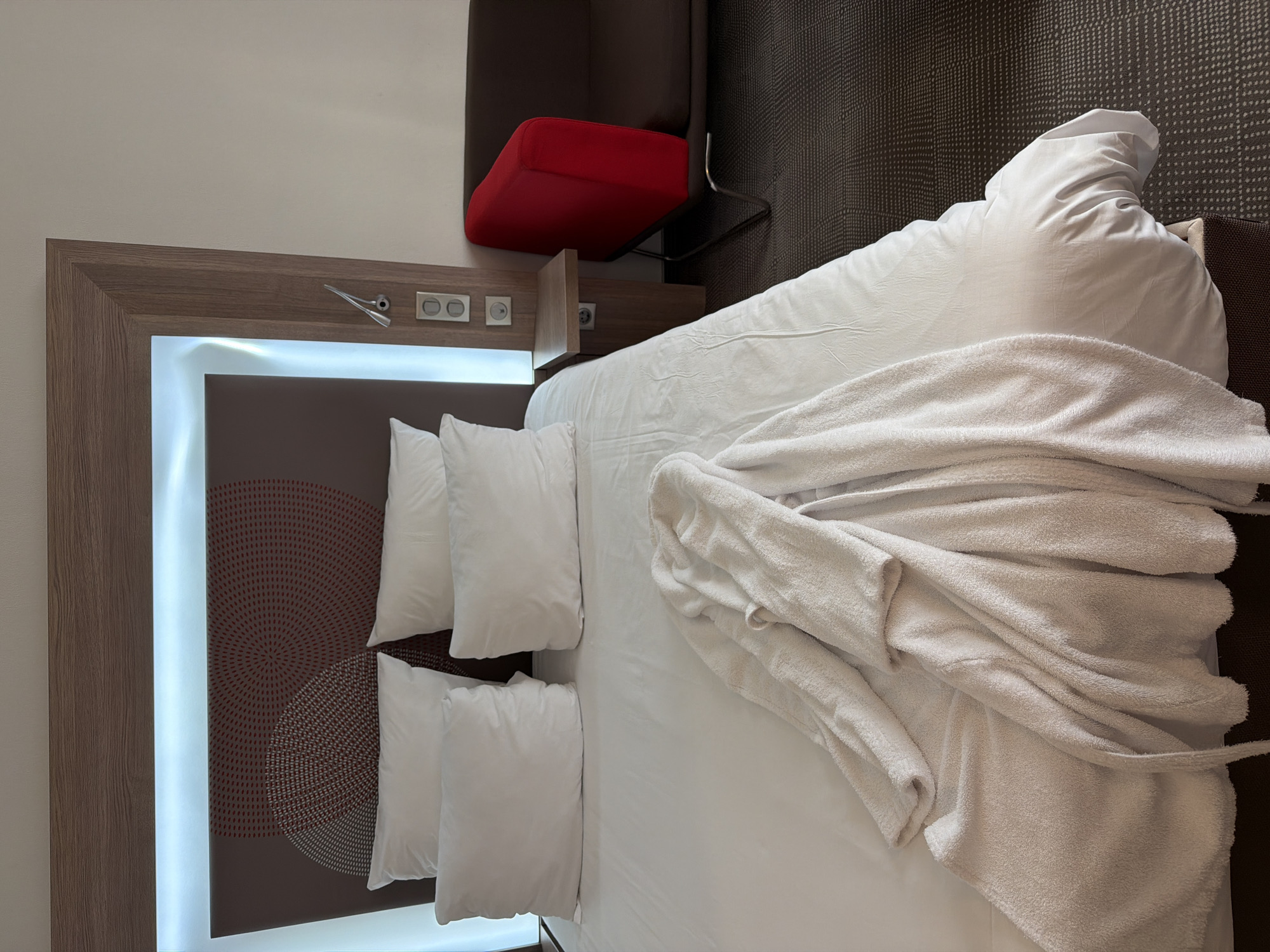 The image depicts a hotel room in Khimki, Russia. A king-sized bed is covered with white linens and several white pillows. A white bathrobe is draped across the bed. To the right of the bed, there is a small wooden bedside table with electrical outlets and a bedside lamp. A dark brown sofa with a bright red accent piece is visible next to the bedside table, situated on a dark patterned carpet. The headboard of the bed is illuminated with white LED lighting and features a circular, patterned design in shades of brown and red. The overall scene is clean and tidy, suggesting a typical hotel guest room.