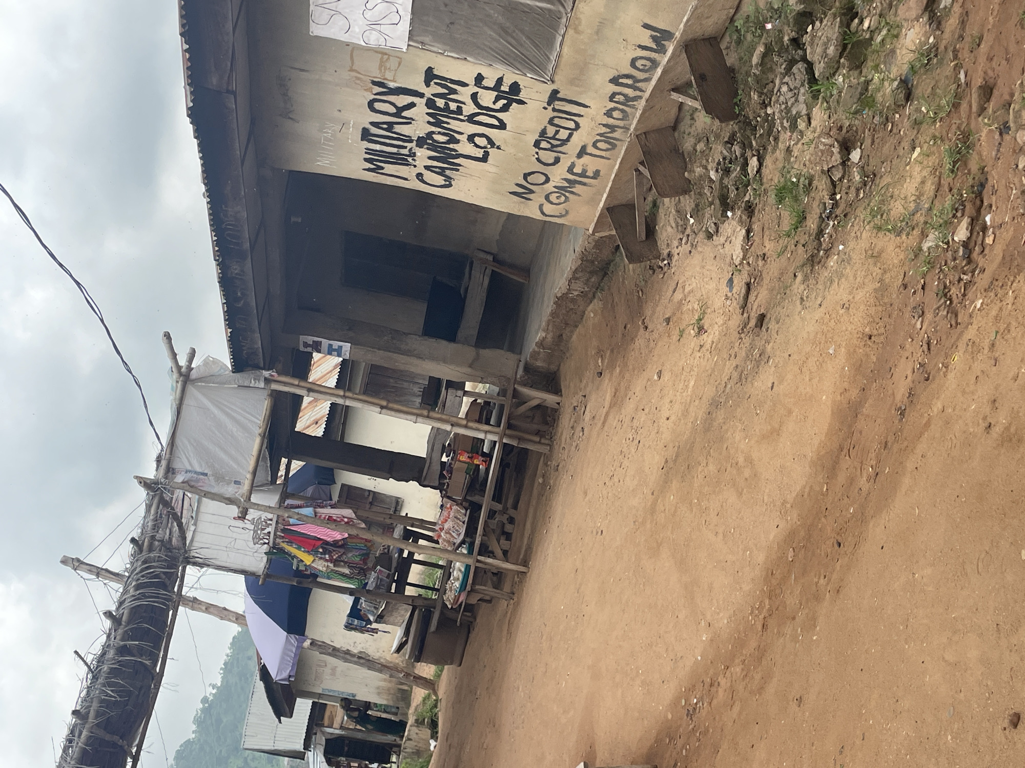 The image shows a dusty street in Lokoja, Nigeria, with a small building on the right side. The building has a corrugated metal roof and is painted a light color. Black lettering on the wall reads "MILITARY CANTONMENT LODGE" and "NO CREDIT COME TOMORROW". A white sign with black lettering is partially visible on the right side of the building, with the word "PASS" clearly legible. In front of the building, there are wooden stands and tables displaying various items, including what appears to be food items and clothing. A few people are visible in the background, one of whom is a woman standing near a stall. The street is unpaved and appears to be made of dirt and gravel, with some scattered rocks and vegetation. The sky is overcast, suggesting a cloudy day. A makeshift structure made of wood and thatch is visible on the left side of the image, possibly a stall or shelter.