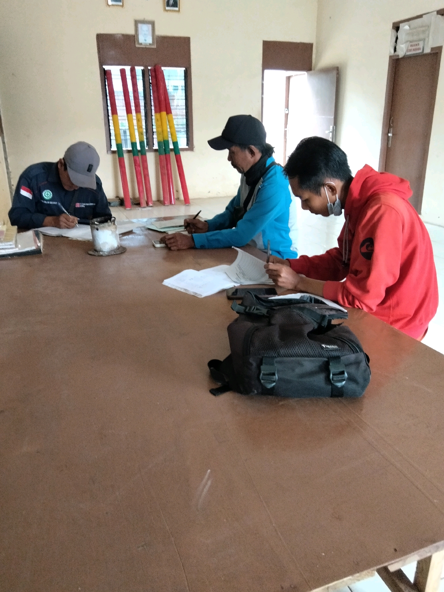 Three individuals are seated at a brown wooden table indoors. On the left, a person wearing a grey baseball cap and a dark jacket with a white and green emblem is writing on a piece of paper. In the center, a person wearing a blue jacket and a black baseball cap is also writing. On the right, a person wearing a red hooded sweatshirt and a grey face mask is writing on a paper. A black backpack is placed on the table in front of the person in the red sweatshirt. In the background, there is a window with several red, yellow, and green poles leaning against it, and two framed pictures on the wall. The environment appears to be a room with light-colored walls and a brown door in the background. The overall scene suggests an indoor gathering or meeting where individuals are engaged in writing activities. The location is Cinta Manis Lama, Indonesia.