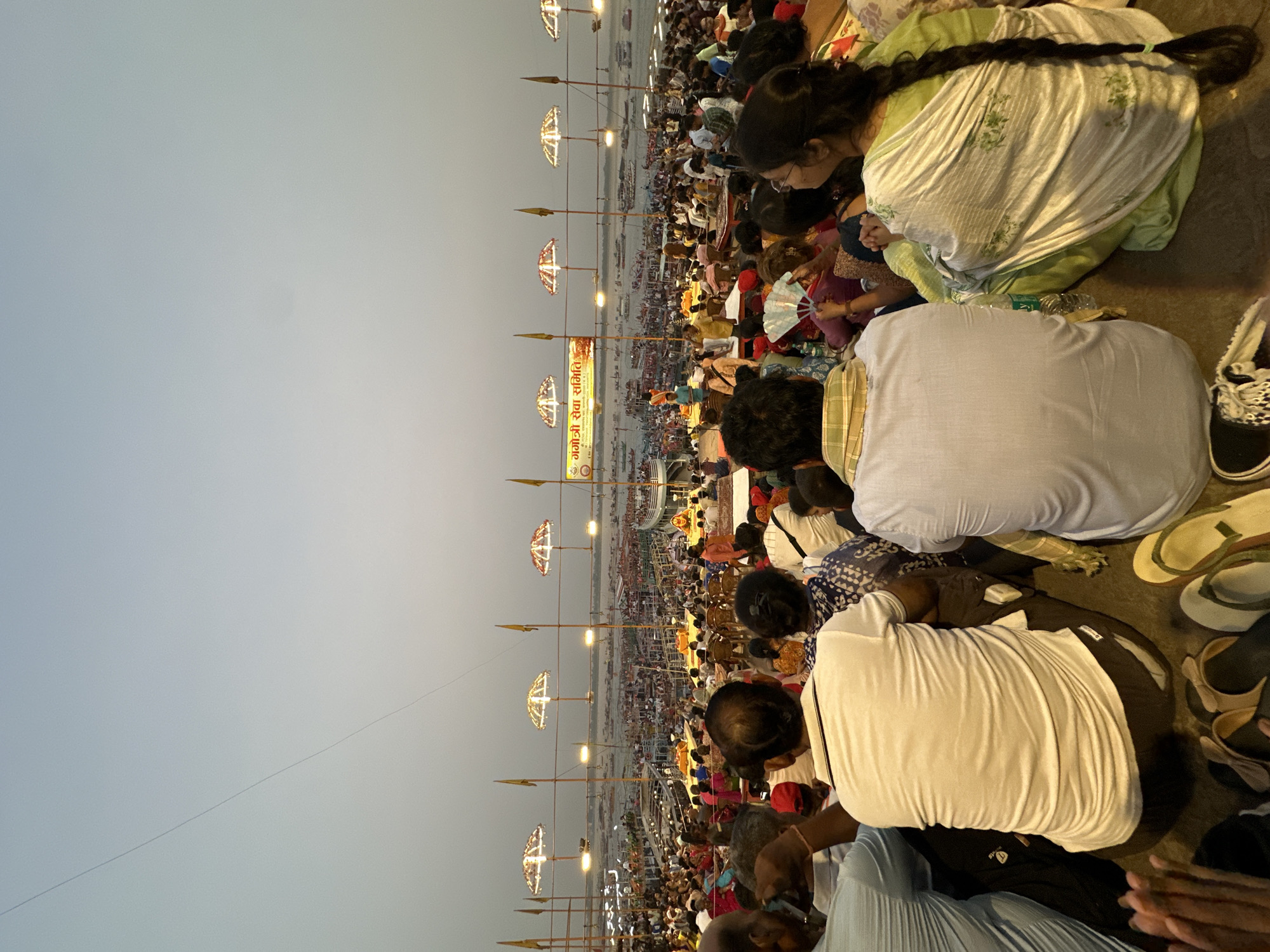 The image shows a large crowd of people gathered near the banks of the Ganges River in Varanasi, India. The photograph is taken from a slightly elevated perspective, looking down upon the crowd. Several people are seated on the ground with their backs to the viewer. There are rows of tall poles with lit decorative structures on top, stretching out towards the river. A banner with Hindi script is visible among the poles. The sky is a pale shade of grey, suggesting it is either dawn or dusk.
