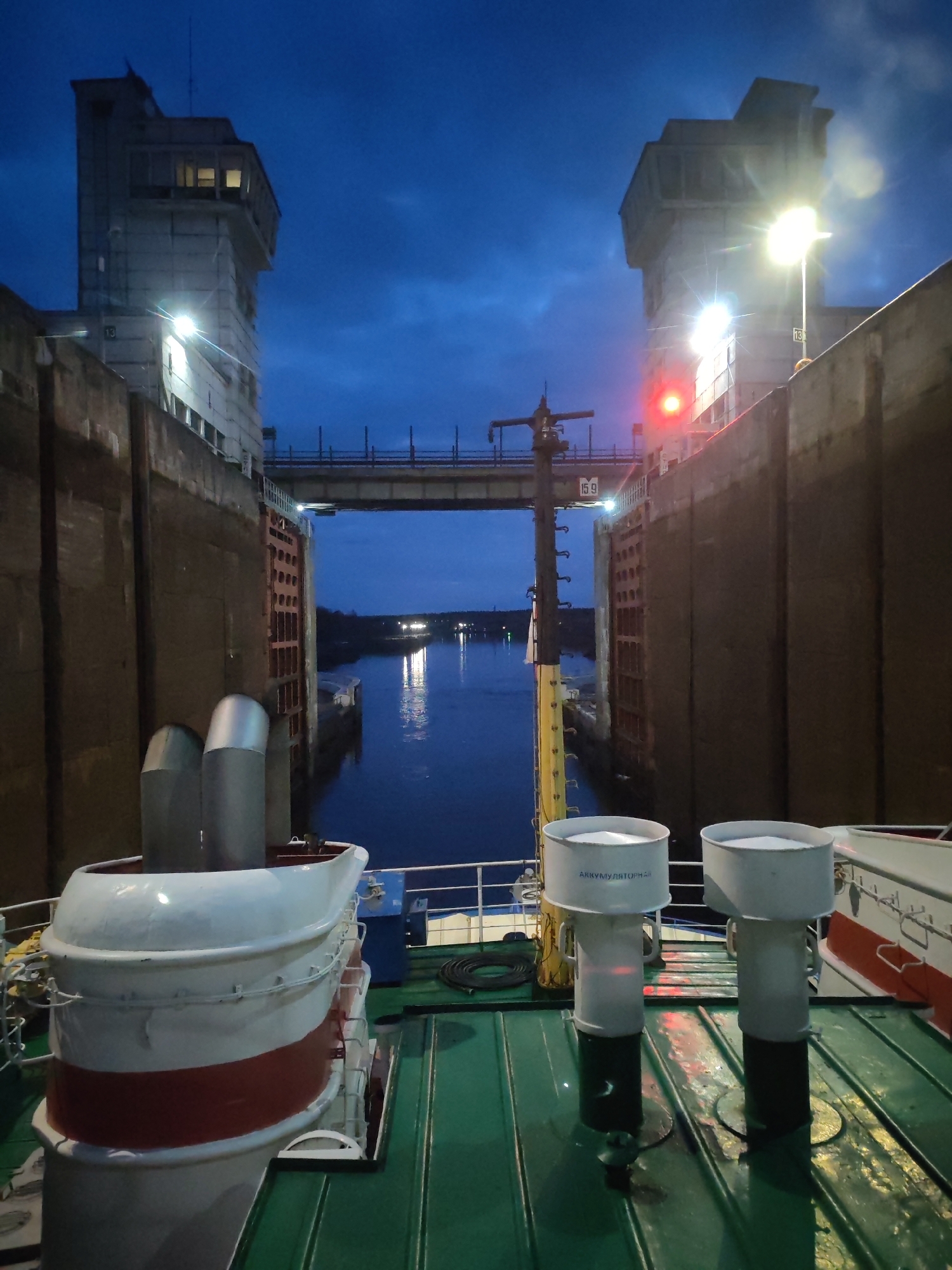 A vessel is depicted inside a large navigation lock chamber during twilight or night. The ship is positioned between massive concrete lock walls, with tall, multi-storied control towers on both sides. The sky above is a dark blue with visible clouds.
Ahead of the vessel, the lock gates are open, revealing a river or canal stretching into the distance, with reflections of distant lights on the calm water. A metal bridge spans the width of the lock chamber, connecting the two sides above the ship's level.
Bright floodlights are mounted on the concrete towers and walls, casting stark illumination on the structures and parts of the ship, contrasting with the dark surroundings. A prominent red signal light is visible on the right-hand control tower/structure.
On the deck of the vessel in the foreground, part of a white and red tender or lifeboat is visible on the left, with two silver cylindrical structures, likely exhaust pipes, rising from it. On the right side of the deck, two white cylindrical vents are present. One of these vents clearly displays the Russian word "АККУМУЛЯТОРНАЯ" (Battery Room).
Notable details include the number "13" visible on the concrete structures of both towers, and the number "15.9" displayed on the bridge structure spanning the lock. A yellow measuring pole or marker is partially visible near the open lock gates. The scene suggests an active waterway infrastructure, likely a river or canal lock system, operating efficiently under artificial light at night.