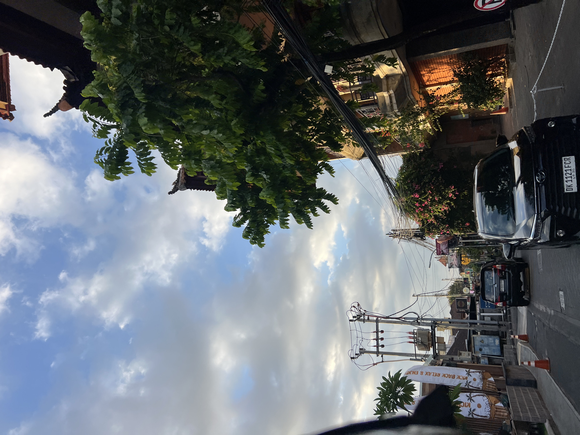 The image depicts a street scene in Kuta, Indonesia, likely during daylight given the clear sky and visible sunlight. The perspective appears to be from inside a vehicle, looking forward. The street is lined with buildings on either side. On the right, a building with traditional Balinese architecture is visible, featuring intricate roof details and balconies. Lush green trees and flowering vines adorn the sides of the buildings, adding to the tropical ambiance.
The street itself is paved and shows signs of urban activity. Several vehicles are present, including a black SUV and a black car, indicating traffic. There are also power lines and utility poles, suggesting the presence of electrical infrastructure. Orange traffic cones are placed along the side of the road, likely for traffic management or temporary obstruction. On the left side of the street, banners with palm tree motifs and text are displayed, possibly advertising local businesses or attractions.
The sky is partly cloudy, with a mix of blue sky and white clouds, illuminated by sunlight that casts shadows and highlights on the buildings and trees. The overall scene conveys a sense of a bustling street in a Southeast Asian city, with a blend of natural elements and urban development.