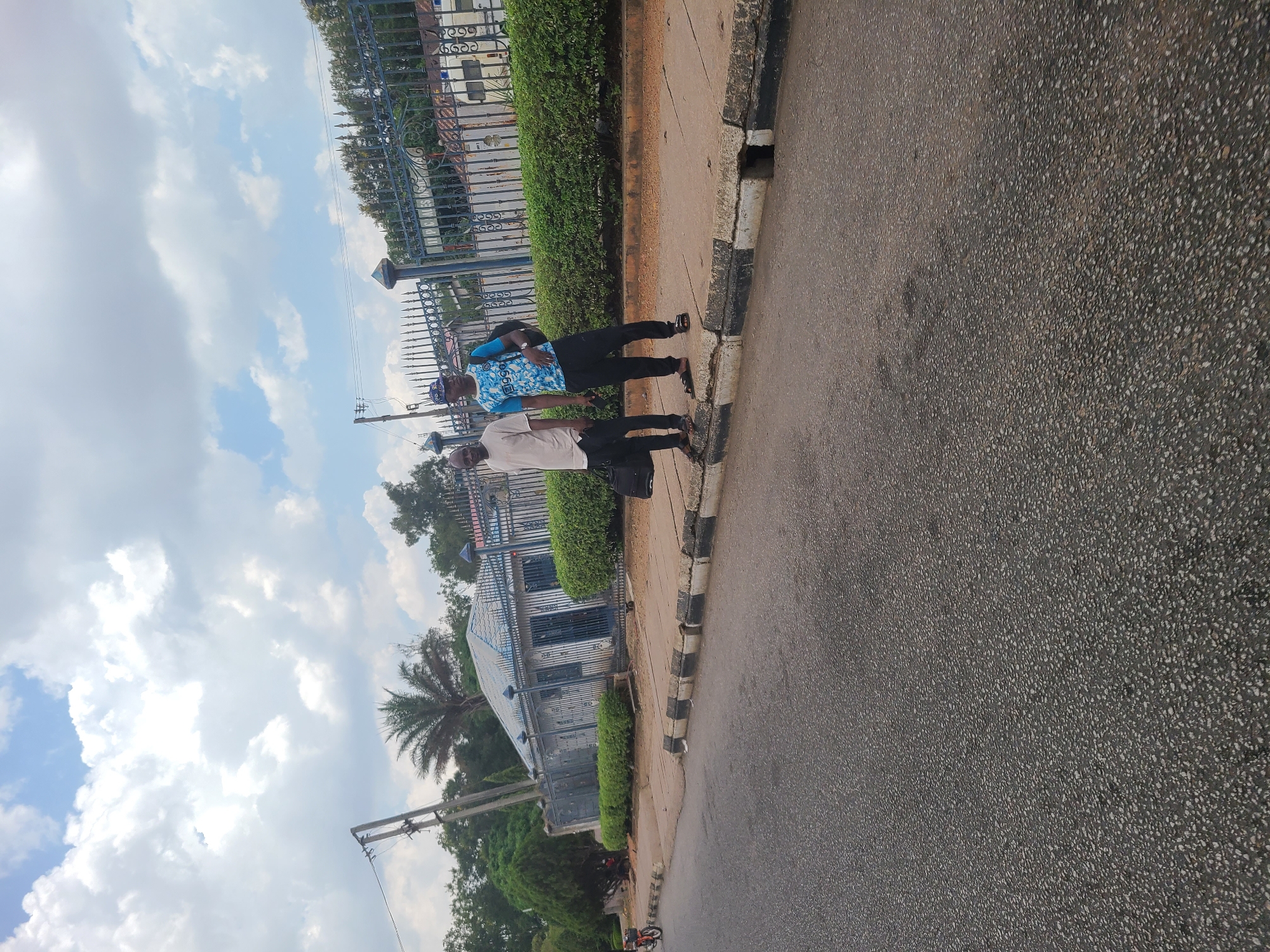 Two men are walking on a sidewalk next to a road. The man in front is wearing a blue patterned shirt, black pants, and sandals. The man behind him is wearing a light-colored shirt, black pants, and sandals. They are walking past a building with a fence and green bushes. The road is asphalt. The sky is blue with white clouds. There is a palm tree in the background. This is in the city of Jos, Nigeria.
