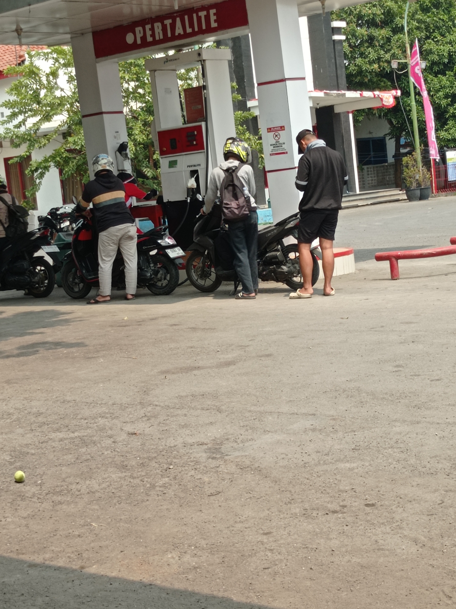 The image captures a scene at a Pertamina gas station in Kramat, Indonesia. In the foreground, the paved ground is visible, with a small yellow ball to the left. Behind it, four individuals are interacting with motorcycles near a gas pump. Two men are standing by their motorcycles, one appearing to be filling his or receiving fuel. Another man is seated on his motorcycle, and a fourth person is also near a motorcycle. The gas station features red and white signage with the word "PERTALITE" prominently displayed. To the right, a red bench and potted plants are visible, along with a pink flag on a bamboo pole. The background shows buildings and trees. The lighting suggests it is daytime, with clear shadows cast on the ground.