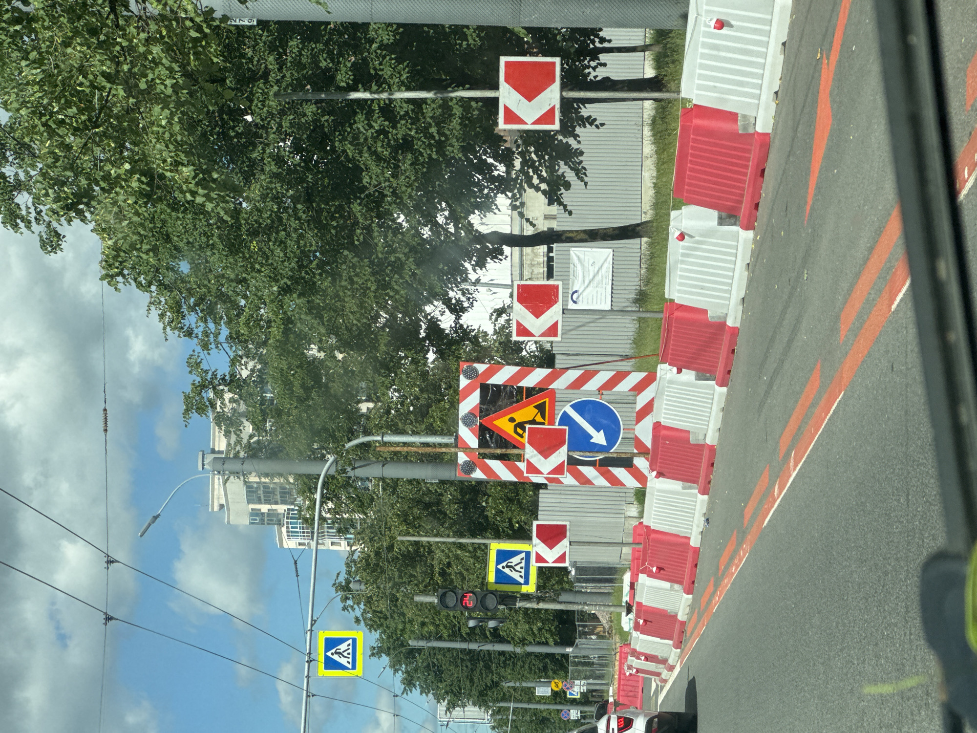 The image depicts a street scene in Saint Petersburg, Russia, characterized by road construction and traffic control measures. A row of red and white traffic barriers delineates a construction zone on the right side of the road, with temporary lane markings in orange indicating traffic flow. Numerous road signs are visible, including pedestrian crossing signs (one yellow and blue, another yellow and white), a traffic light showing "24", a large rectangular sign with diagonal red and white stripes, a yellow triangular sign depicting a construction worker, a red and white sign with a left-pointing arrow, and a blue circular sign with a white arrow indicating a mandatory left turn. Overhead power lines crisscross the sky, which is partly cloudy. In the background, a multi-story building and lush green trees are present. The perspective is from inside a vehicle, looking forward.