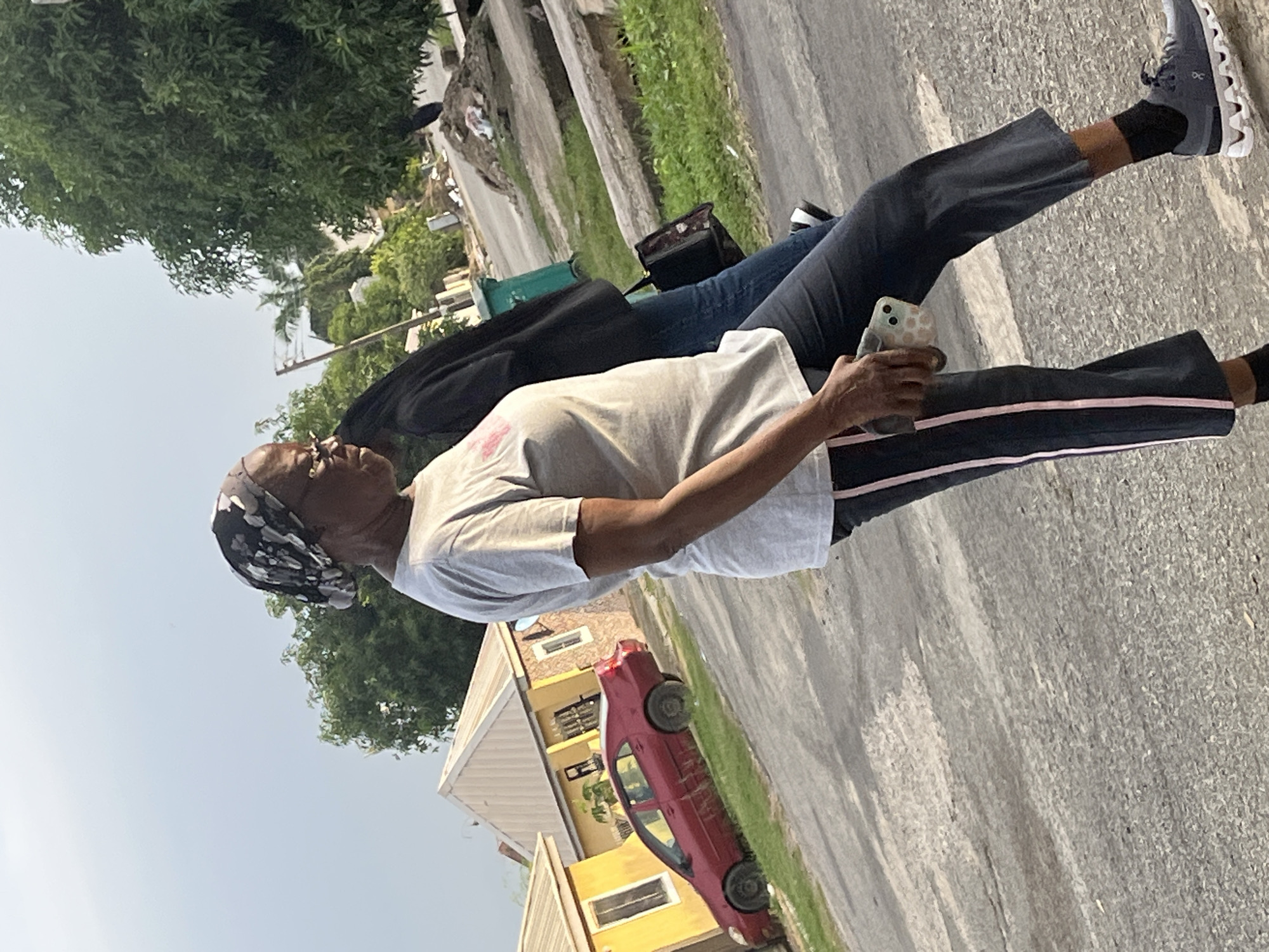 The image depicts an outdoor scene in Abuja, Nigeria, featuring two individuals walking on a paved street lined with residential buildings and vegetation. The foreground is dominated by a woman walking in profile towards the right of the frame. She is wearing a light grey t-shirt, dark trousers with a pink stripe down the side, black socks, and grey athletic shoes. A patterned headscarf covers her hair, and she wears glasses. In her right hand, she holds a smartphone with a patterned case. Behind her, another person is partially visible, wearing a dark jacket and blue jeans, carrying a patterned bag. In the background, a red car is parked on the grass beside a yellow building. The environment suggests a residential area with houses, trees, and a green trash bin visible. The sky is overcast.