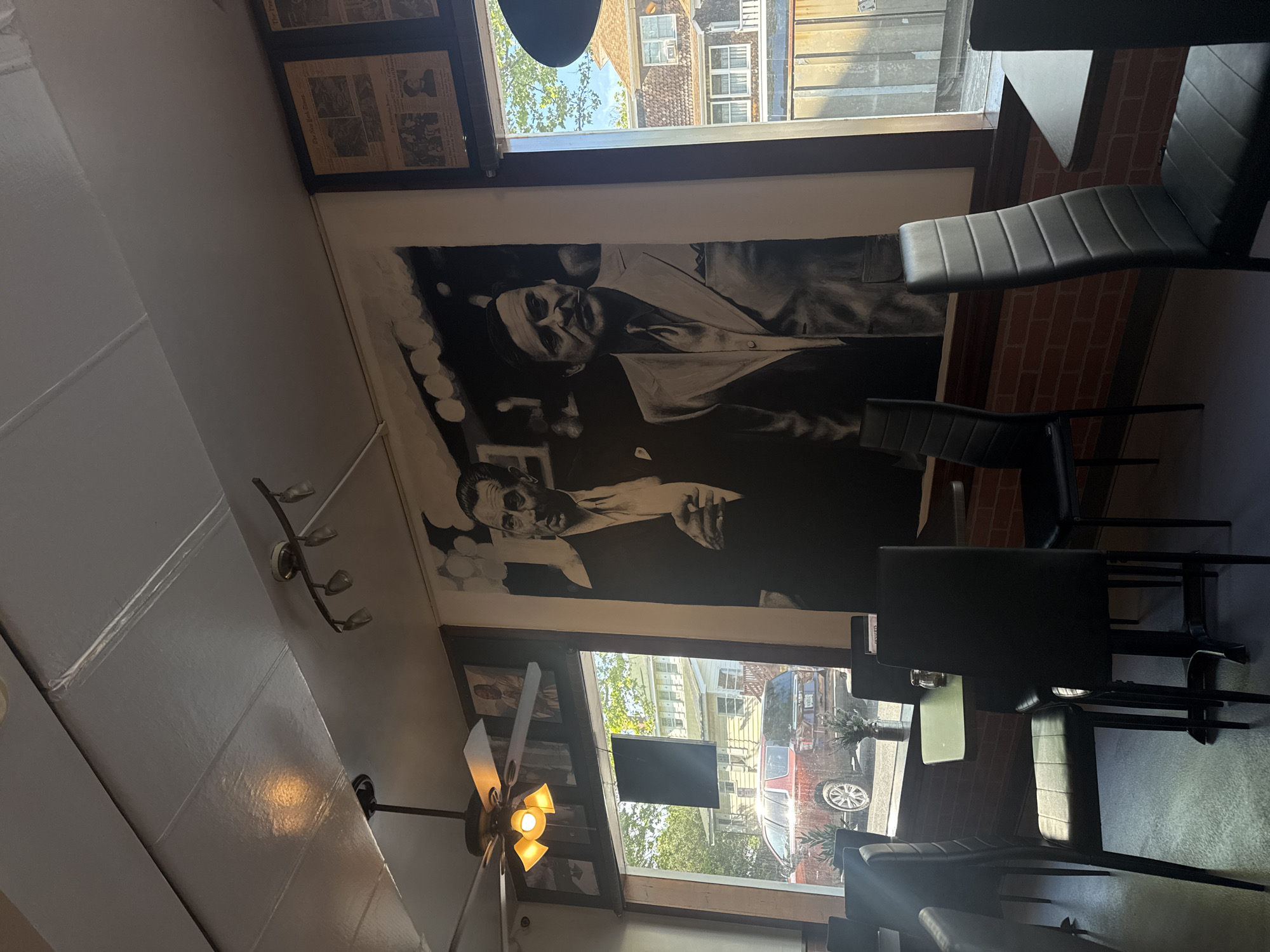 The image shows an interior scene, likely a restaurant, possibly in Ocean City, United States of America. The orientation of the image is turned ninety degrees clockwise. A black and white portrait of two men dominates the top wall space above a window. Seating, consisting of black chairs and tables, appears to be arranged near the window. A ceiling fan is visible near the bottom window. The view outside the window includes buildings and cars. Another window shows newspapers or other framed content.