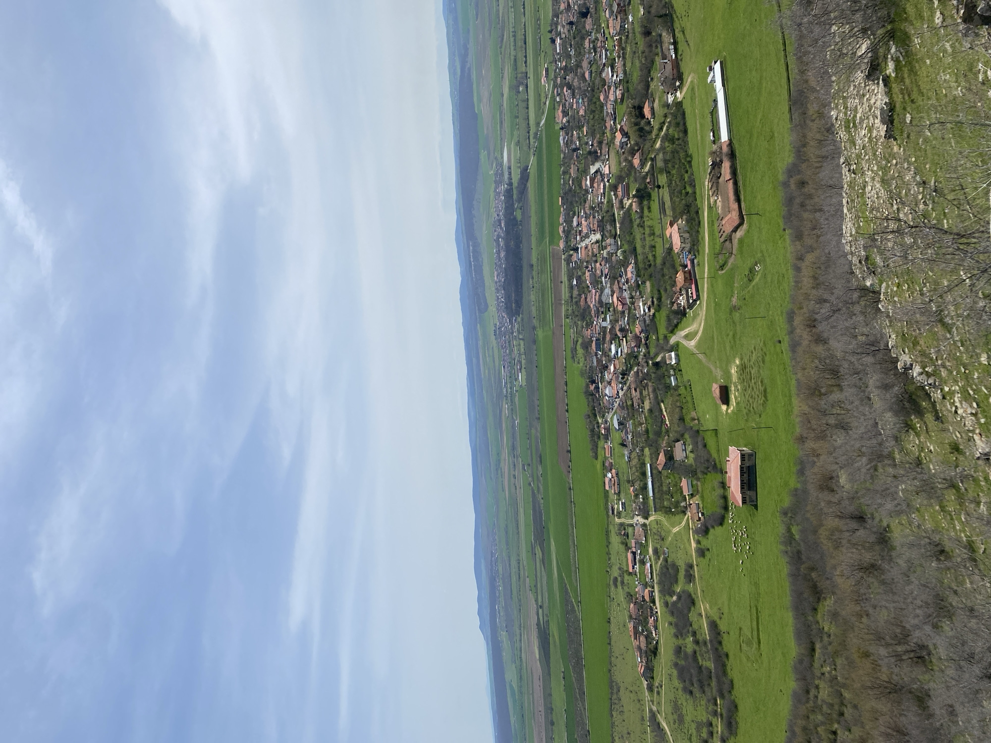The image shows an aerial view of Goritsa, Bulgaria, a rural village nestled between green fields and a rocky ridge. A cluster of houses with red tile roofs forms the core of the village, extending along a dirt road. A long, white industrial building stands out against the green landscape near the ridge. Patches of forest and shrubbery are visible along the edge of the rocky area, while open green fields stretch towards a distant horizon. The sky is partially covered with white, wispy clouds. A flock of sheep is located in an open green field, some distance away from a building with a red roof.