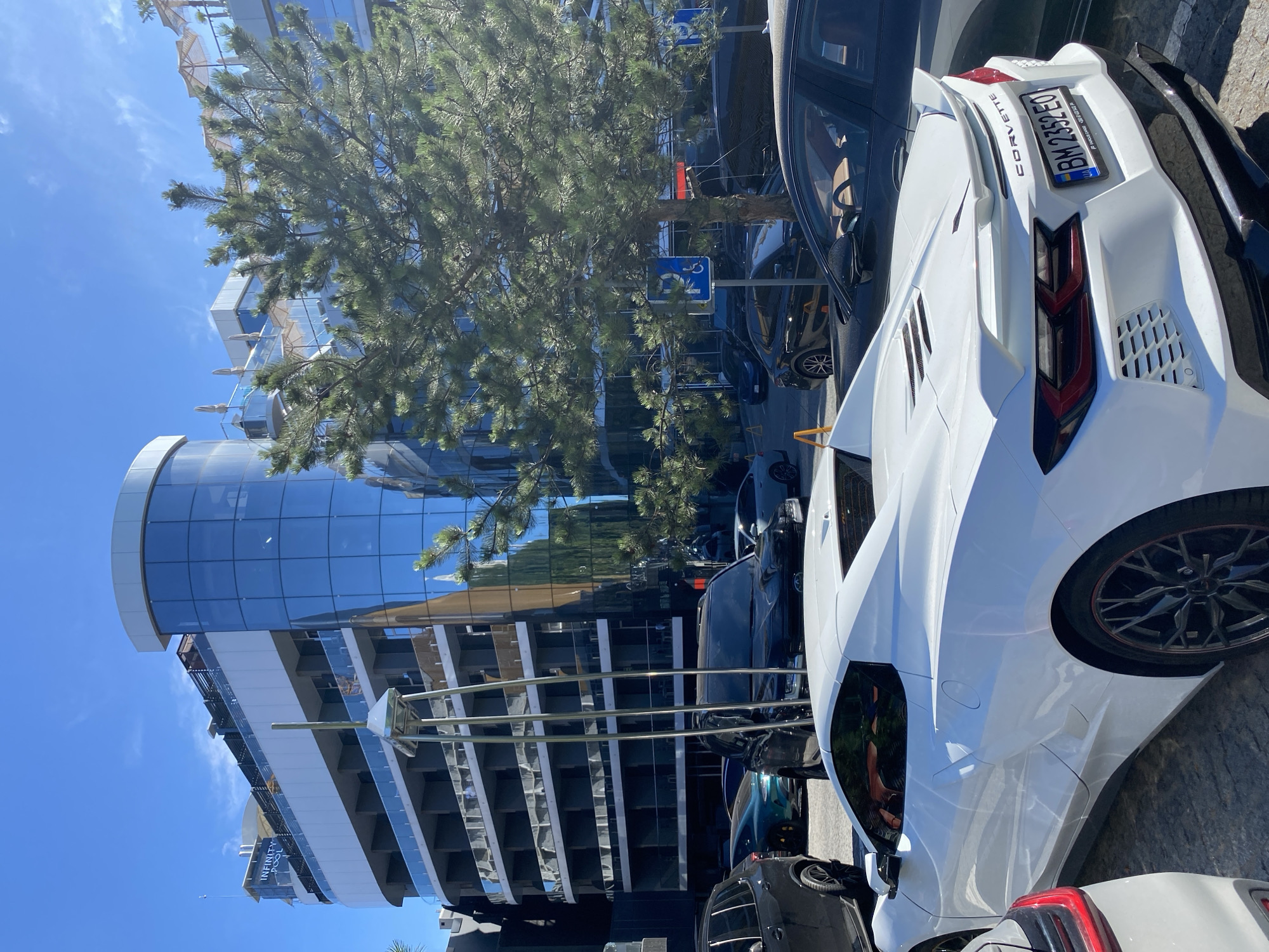 A white Chevrolet Corvette is parked on a cobblestone surface in front of a modern building with a curved glass facade. The building has multiple floors with balconies and is topped with a rooftop area featuring umbrellas and lounge chairs. A large evergreen tree with dense foliage is positioned in the foreground, partially obscuring the view of the building. Several other vehicles, including dark-colored SUVs and sedans, are visible parked in the background. The sky is clear and blue, indicating a sunny day. A blue parking sign with a wheelchair symbol is also present near the tree. The license plate of the Corvette is BM 2352 EO.