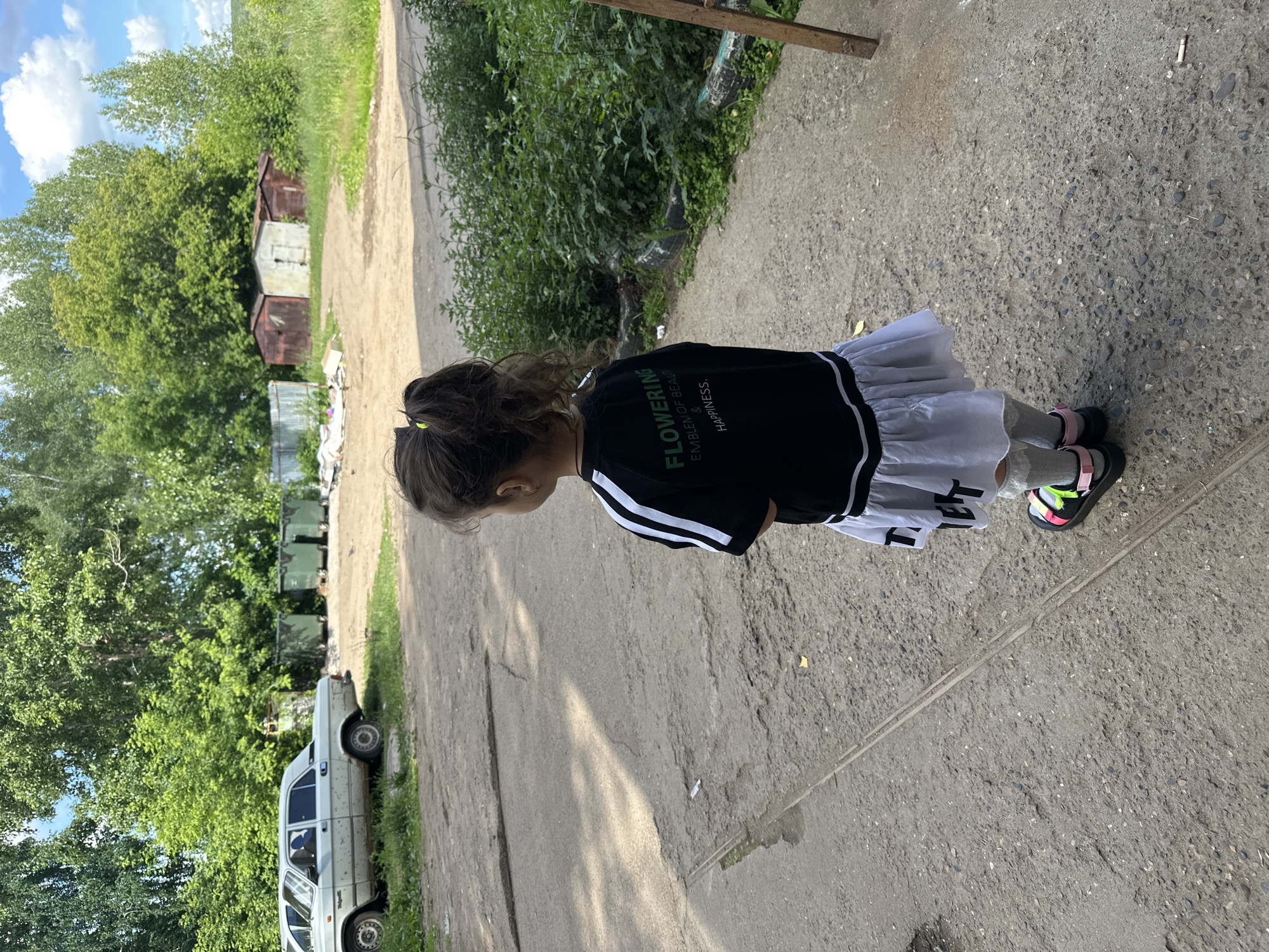 A young girl stands on a paved path in an outdoor setting. She is wearing a black t-shirt with white stripes on the sleeves and a white skirt. Her hair is tied back in a ponytail. She is wearing sandals on her feet.
In the background, there is a white car parked on the side of the path, and further back, a line of green dumpsters. Behind the dumpsters and the car, there are numerous trees and lush green foliage. Further in the distance, there are a couple of old, rusted sheds. The sky is blue with some white clouds, suggesting a fair weather day. The light indicates it is daytime.
The image is taken from a high angle, looking down at the girl. The path appears to be a rural or semi-industrial area, given the presence of the sheds and dumpsters. There is text visible on the girl's t-shirt: "FLOWERING," "EMBLEM OF BEAUTY," and "& HAPPINESS." The overall impression is a casual outdoor scene.