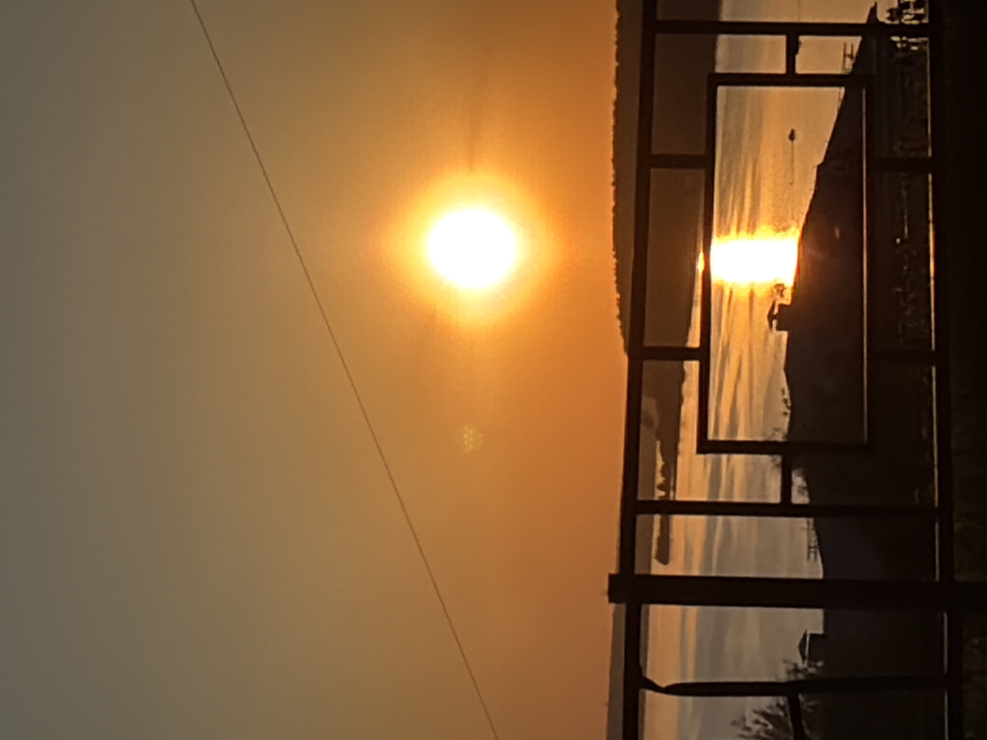 The image captures a sunset viewed through a dark metal framework, possibly a fence or railing. The sun is a bright, spherical shape in the upper part of the image, emitting a radiant glow. Its reflection shimmers on the surface of the water visible through the framework, suggesting a river or lake. Land is also visible in the spaces between the metal bars. A power line crosses from the top left, slightly disrupting the sky. The scene appears to be in or near Izhevsk, Russia, given the request's context. However, no specific landmarks or other identifying features confirm its exact location within the city.