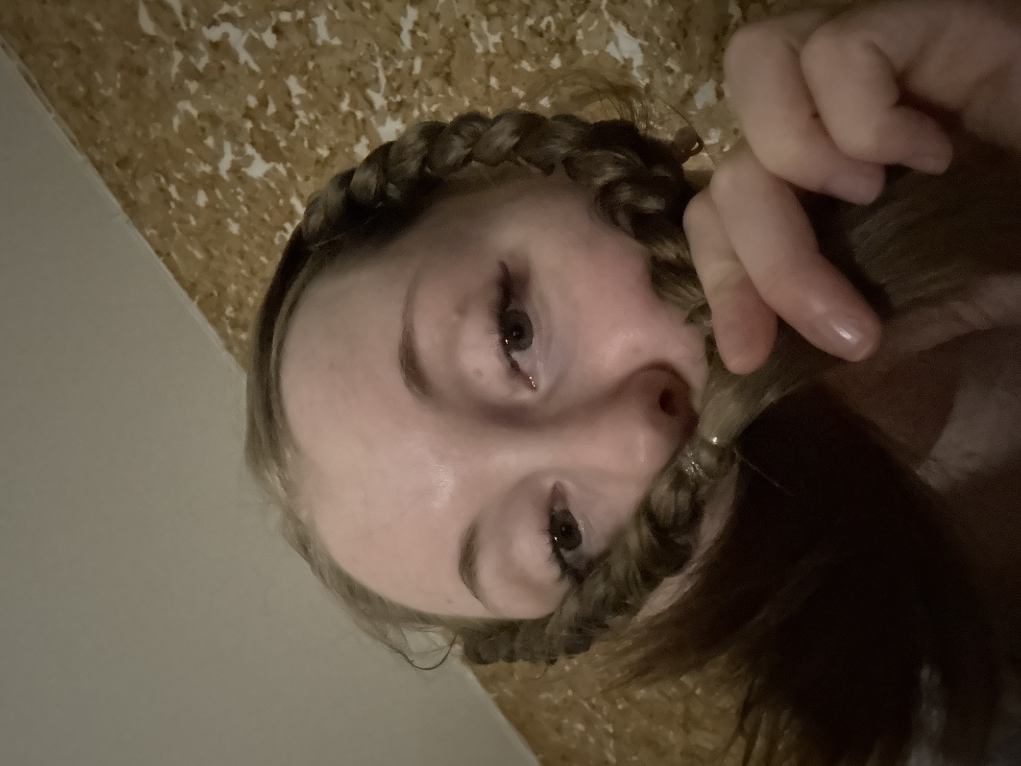 The image captures a close-up, upward-angled portrait of a young person, likely female, with fair skin and light brown hair. The photograph appears rotated 90 degrees clockwise, showing the individual either lying down or looking up at the camera.
The central activity involves the person playfully holding a section of their braided hair across their face, just below the nose, creating a "braid mustache" effect. One hand, with fingers visible and nails appearing to have clear polish, holds this braid. A small, shiny, possibly glittery, detail or accessory is noticeable on the braid near the nose.
The person's eyes are open and looking directly at the camera. They have dark pupils and appear to have mascara or eyeliner applied. The skin shows a slight sheen under what seems to be indoor lighting. The hair is styled with prominent braids, possibly a crown braid or similar, wrapping around the head.
The background is divided into two sections. On the lower left (if rotated to standard orientation), there is a plain, light-colored wall. The remainder of the background is a textured, mottled surface in shades of brown, beige, and white, resembling cork board, patterned wallpaper, or an acoustic panel.
The scene conveys a casual and lighthearted mood. There is no visible text. The indoor setting suggests artificial light.
