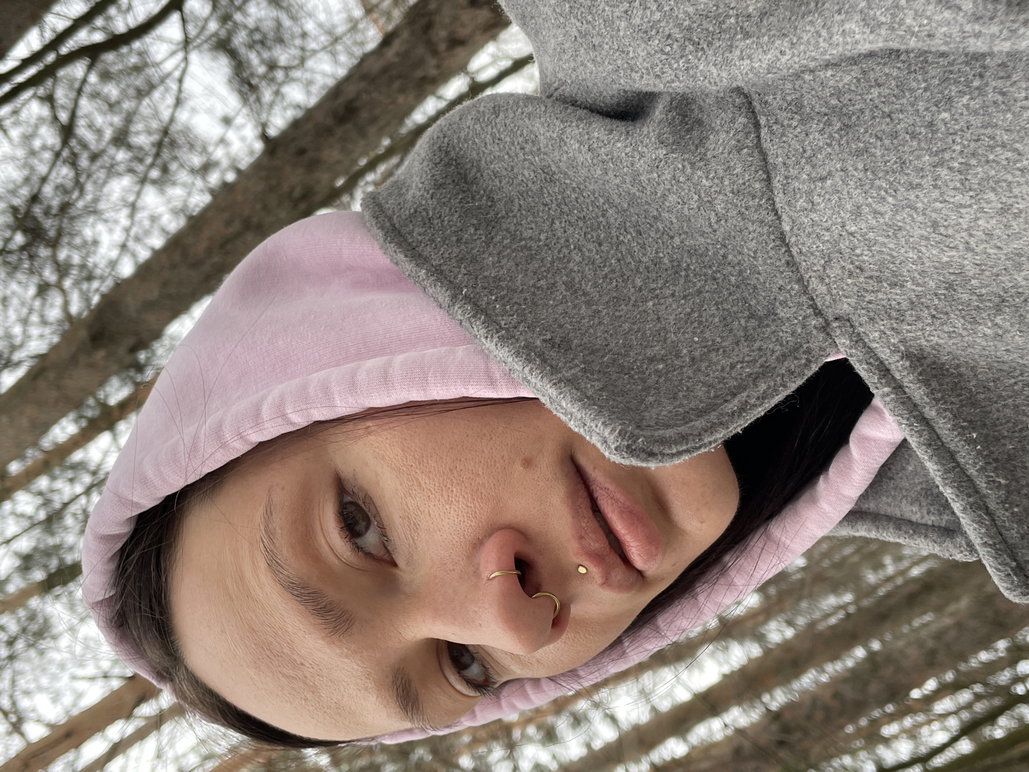 A close-up, upward-tilted, angled shot depicts a person outdoors, likely in a wooded area, possibly near Izhevsk, Russia. The individual has light skin, dark hair partially concealed by a light pink hooded sweatshirt, and a gray wool coat. The person's face is prominent, featuring brown eyes, a gold nose ring, and a lip piercing. The background shows out-of-focus tree trunks and branches.