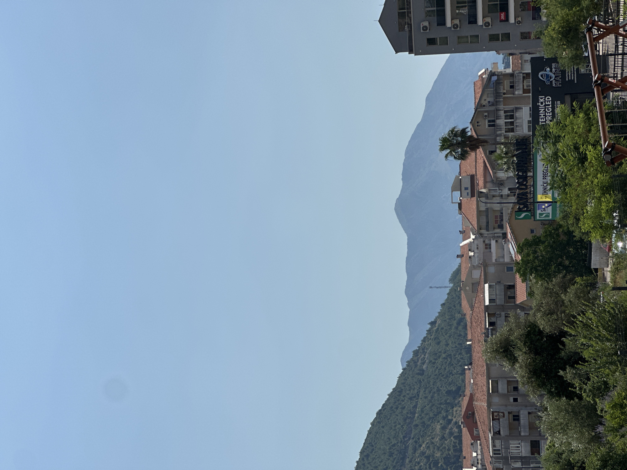 The image depicts a view of buildings and a mountain range under a clear blue sky, suggesting daytime and fair weather. The orientation of the image is portrait, rotated 90 degrees clockwise.
In the foreground, on the right side of the frame, a row of buildings with red-tiled roofs is visible, nestled against a lush green, tree-covered mountainside. These buildings appear to be residential or commercial, with multiple windows and balconies. A tall palm tree stands out among the greenery, adding a tropical element to the scene. To the far right, a section of a dark, metal structure, possibly playground equipment or scaffolding, is partially visible.
Further back, a larger, multi-story building with a grey facade is seen. This building has numerous windows and air conditioning units mounted on its exterior. A prominent black sign with white text is attached to one of the buildings, reading "TEHNIČKI PREGLED MAINE" and below it, "PREGLED VOZILA". Another smaller green sign with white text is also visible.
The backdrop is dominated by a majestic, hazy blue mountain range that recedes into the distance. The texture of the mountains suggests a rugged, natural landscape.
The scene appears to be a calm, everyday view of a town or village situated at the foot of mountains, possibly in a coastal region given the presence of palm trees. There are no people visible in the frame, and the overall impression is one of quiet observation of the urban and natural environment. The location context is Donji Pobori, Montenegro.