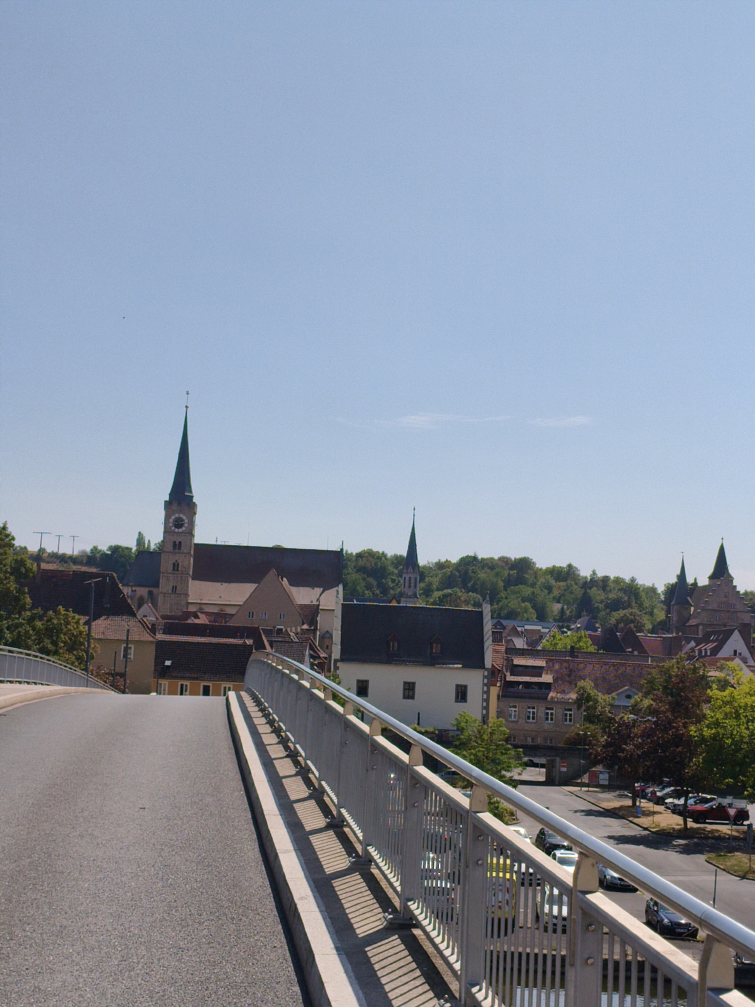 This is a vertical, medium-shot photograph taken from a bridge overlooking the city of Ochsenfurt, Germany. The foreground features the asphalt surface of the bridge, curving to the left, with a metal railing running along its edge. The railing is composed of vertical bars and horizontal beams, casting shadows on the bridge's lower ledge. Beyond the bridge, a dense collection of buildings with terracotta-tiled roofs dominates the middle ground. A prominent church steeple, featuring a clock face, rises above the rooftops, with a tall, slender spire. Further into the background, lush green trees form a backdrop to the cityscape, with more church spires visible in the distance. The sky above is a clear, pale blue. A few cars are parked and driving on a street visible to the right of the frame, next to the buildings.